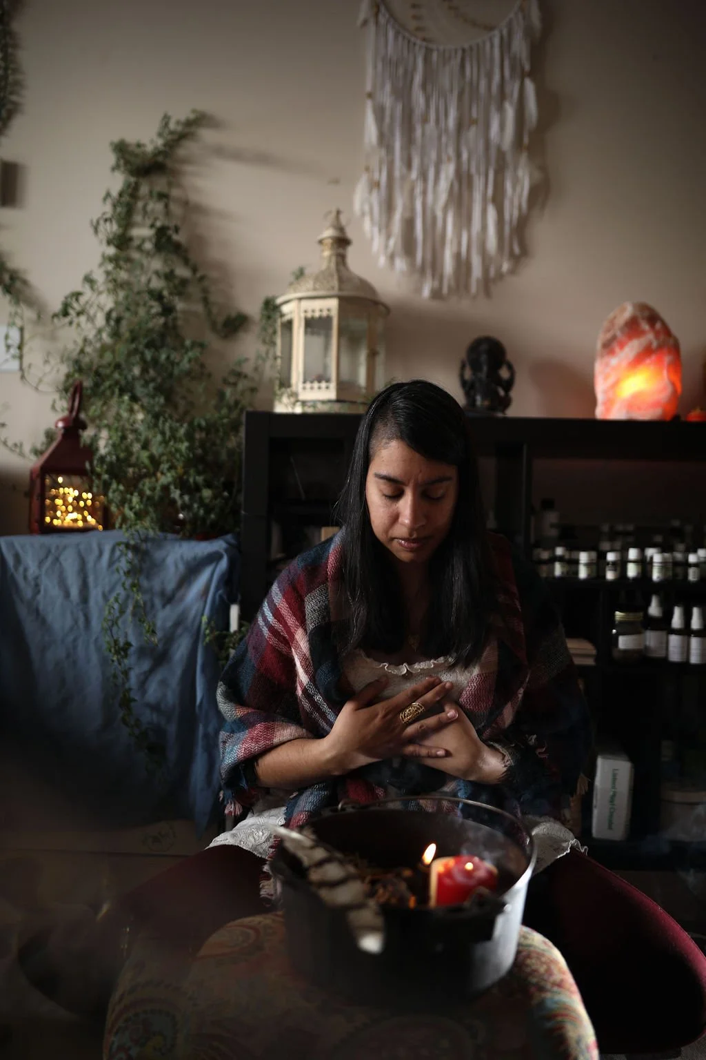 Reverend Clarissa is sitting in prayer over a sacred bowl and candle, her hands are at her heart and she is in her work sanctuary with sacred objects all around her.