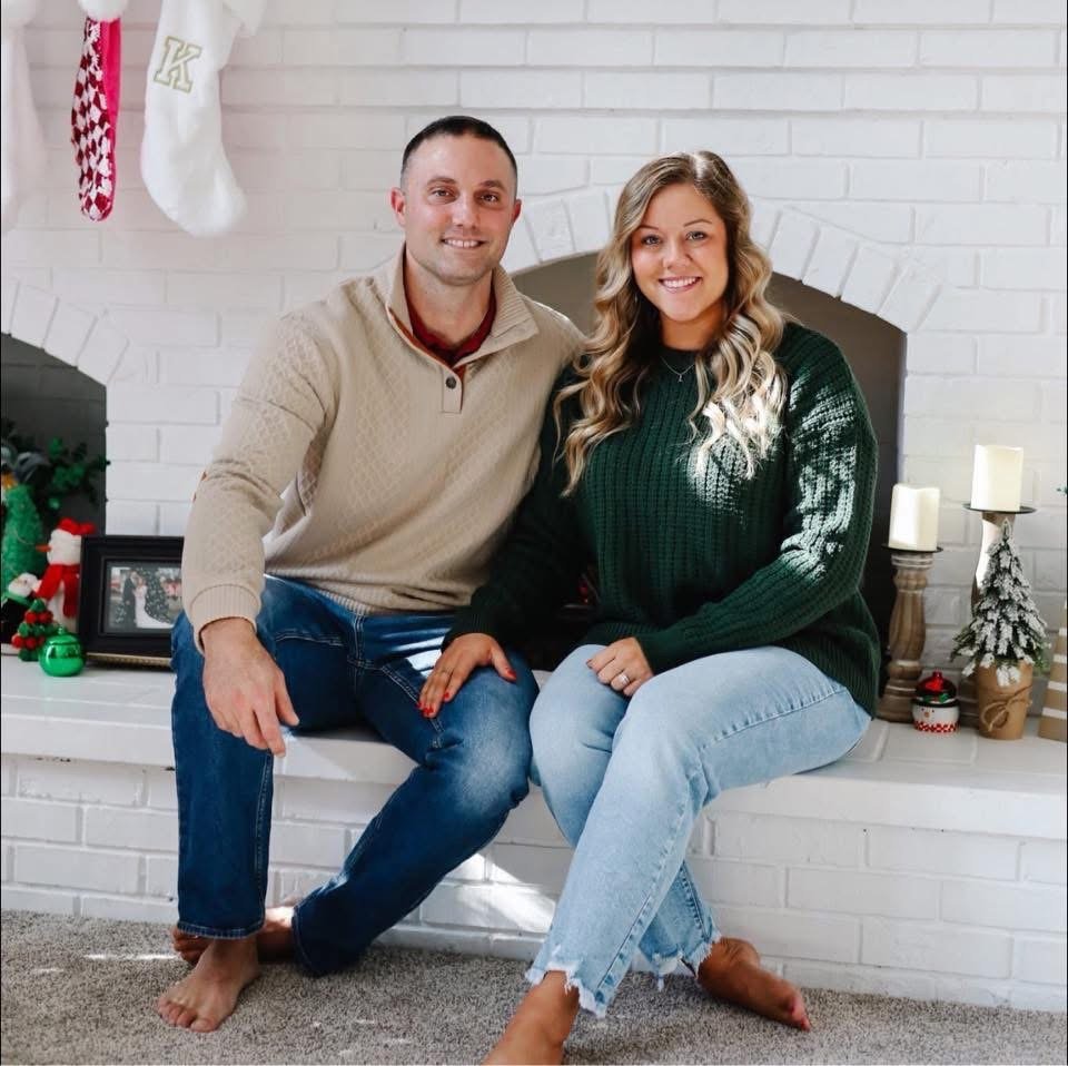 A smiling couple sitting on a white brick fireplace hearth, decorated for Christmas with stockings, Christmas themed figurines, candles, and small trees.