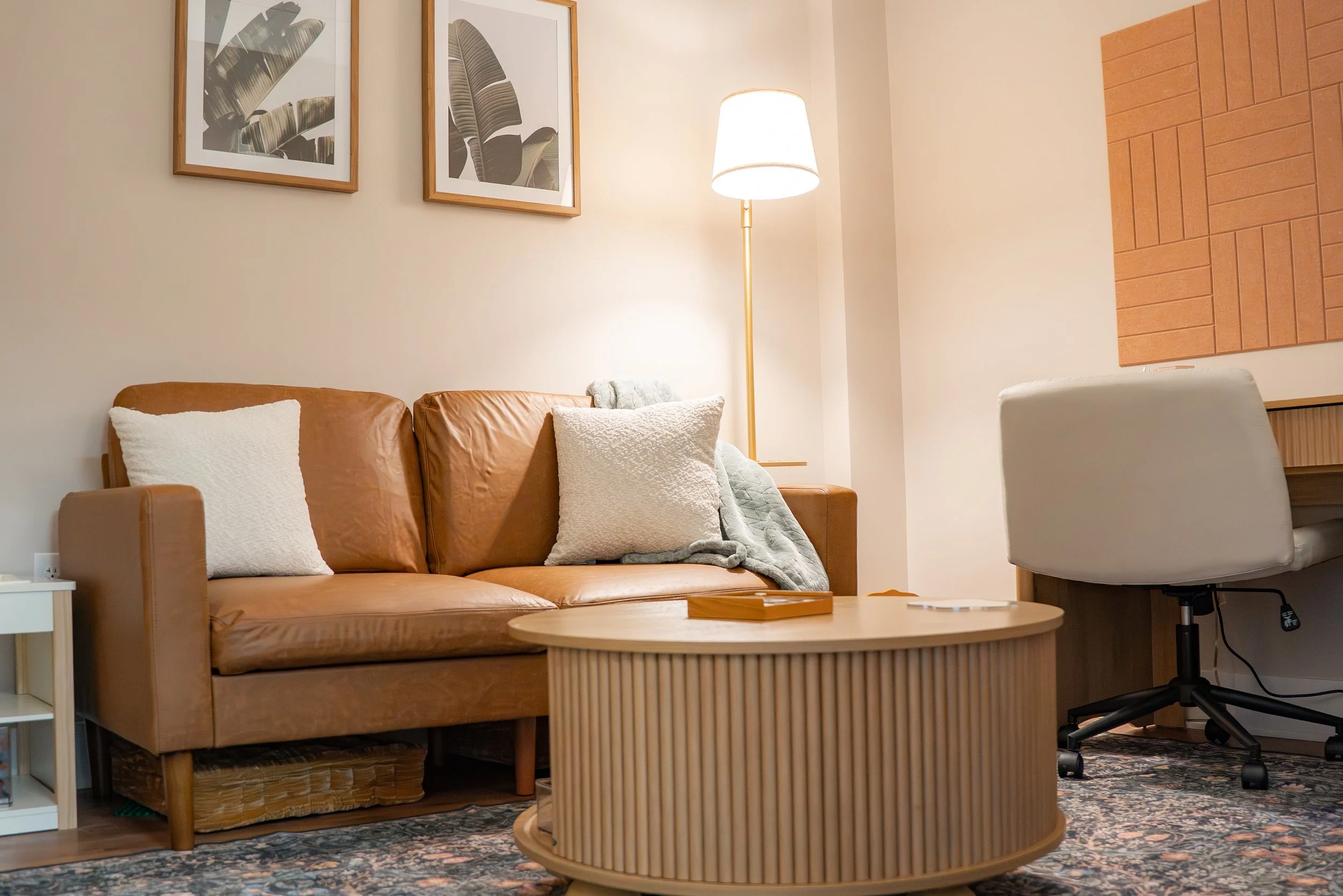 Living room with a tan leather sofa decorated with pillows, a wooden oval coffee table, a white desk chair, wall art featuring leaf prints, a tall floor lamp, a small white side table, and a patterned rug.