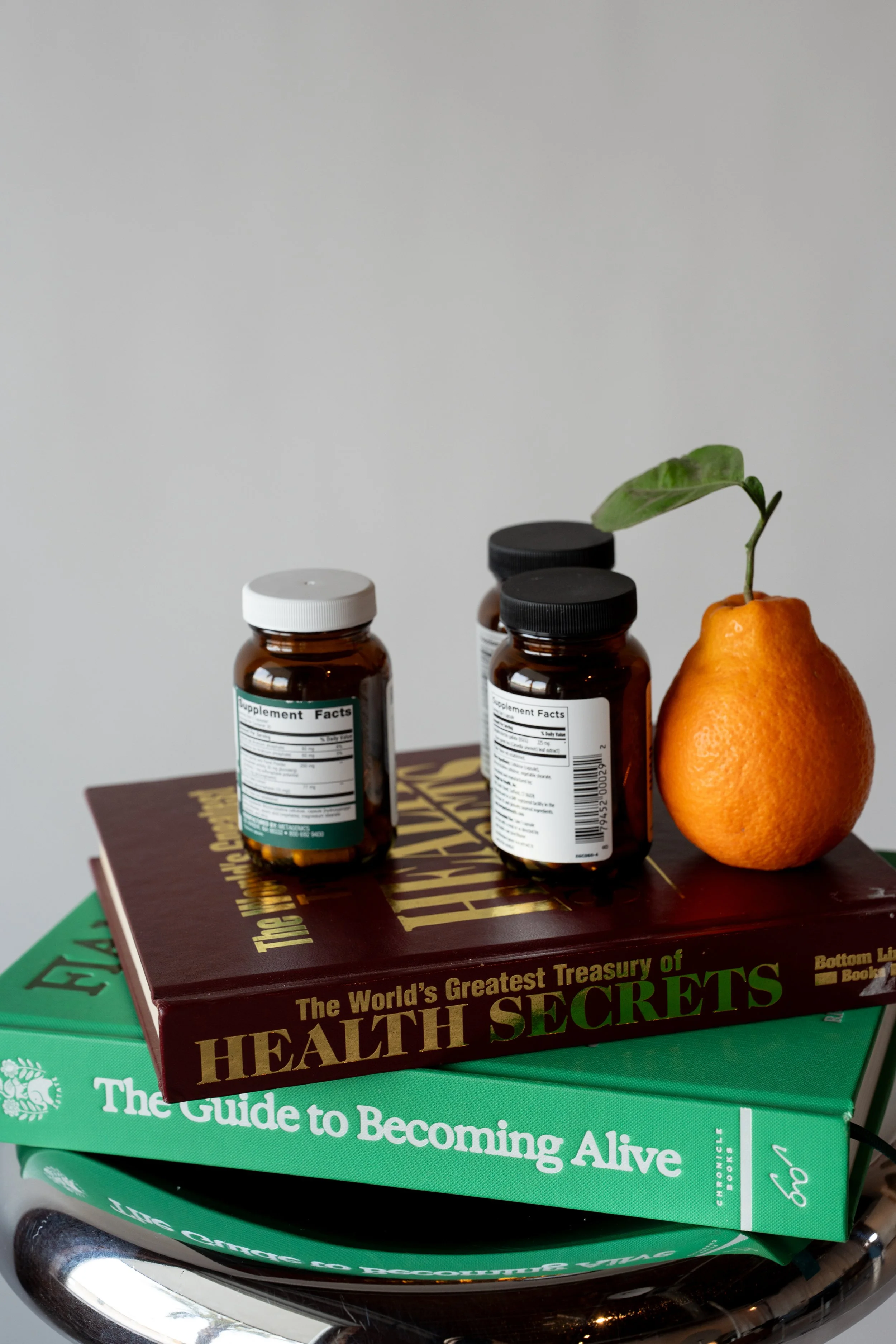 Three supplement bottles, a pear with a leaf, and two books titled 'The World's Greatest Treasury of Health Secrets' and 'The Guide to Becoming Alive' stacked on a metallic surface.