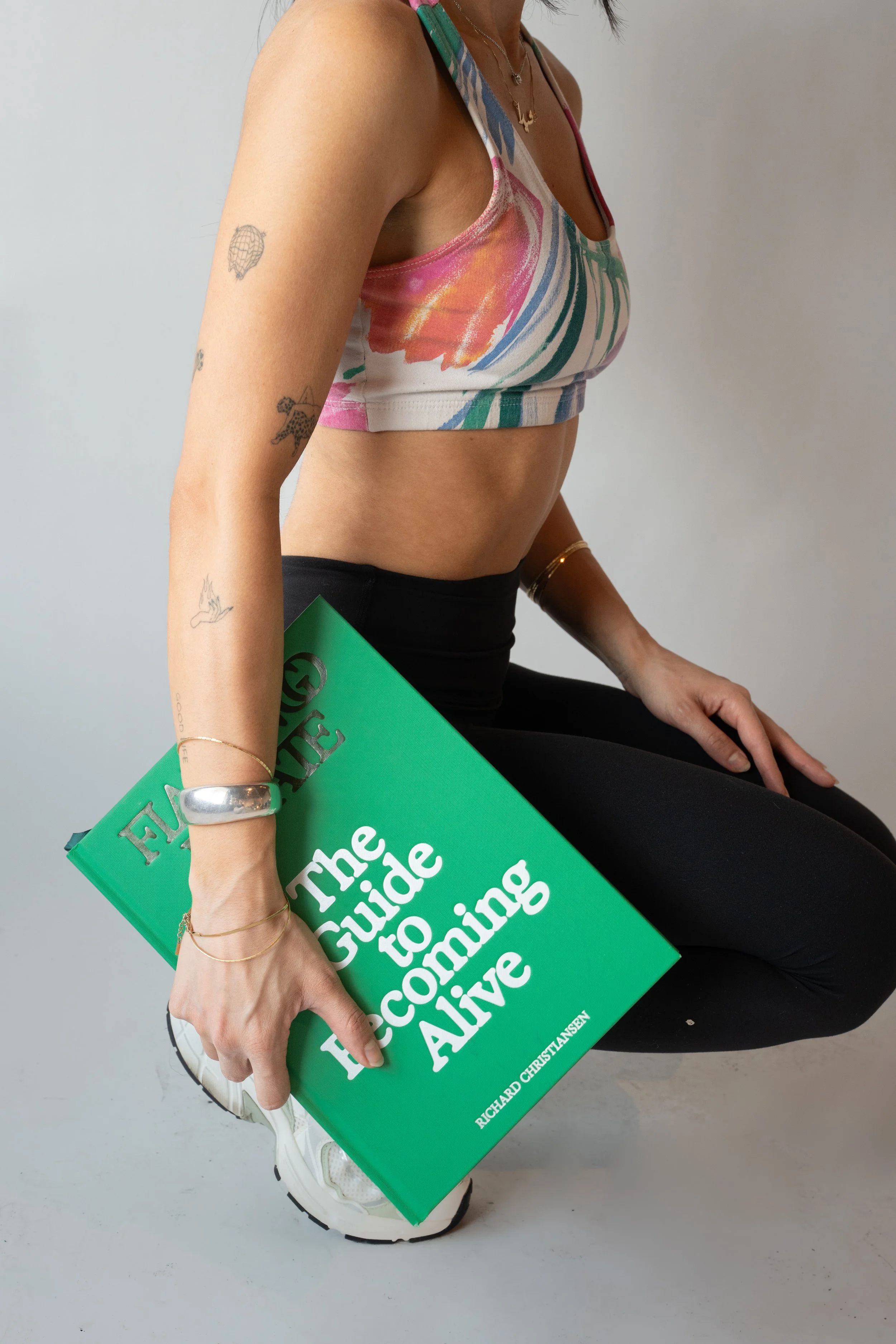A person holding a green book titled 'The Guide to Becoming Fearless' by Richard Christy Jansen. They are sitting on the floor, wearing a colorful sports bra and black leggings, with tattoos on their left arm and gold jewelry.