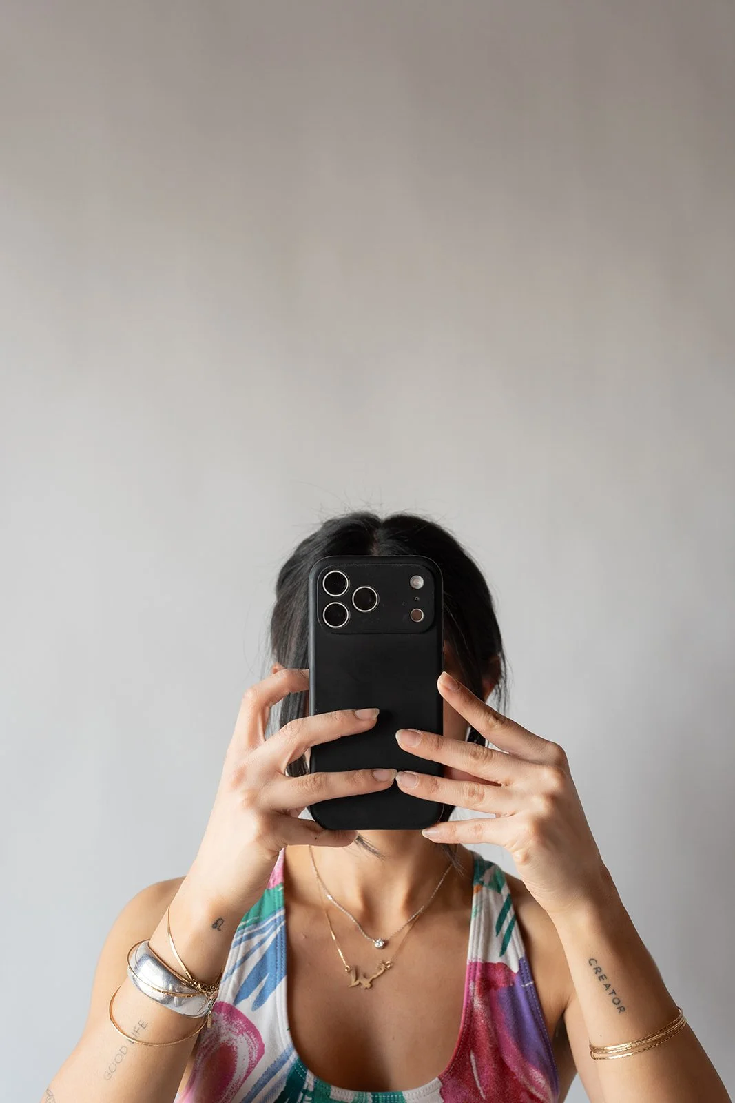 A woman taking a selfie with a smartphone in front of a plain light-colored wall, wearing a colorful sleeveless top, jewelry, and bracelets.