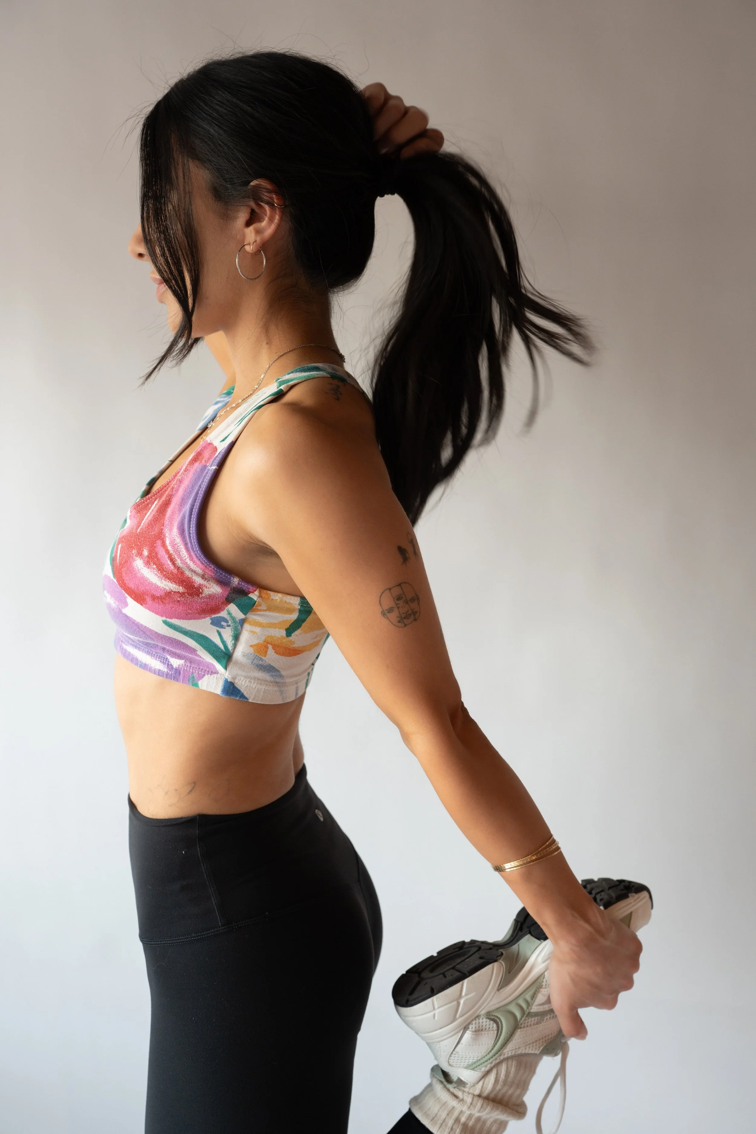 Woman holding her foot behind her back, stretching, wearing athletic clothing, with tattoos and jewelry, in front of a plain neutral background.