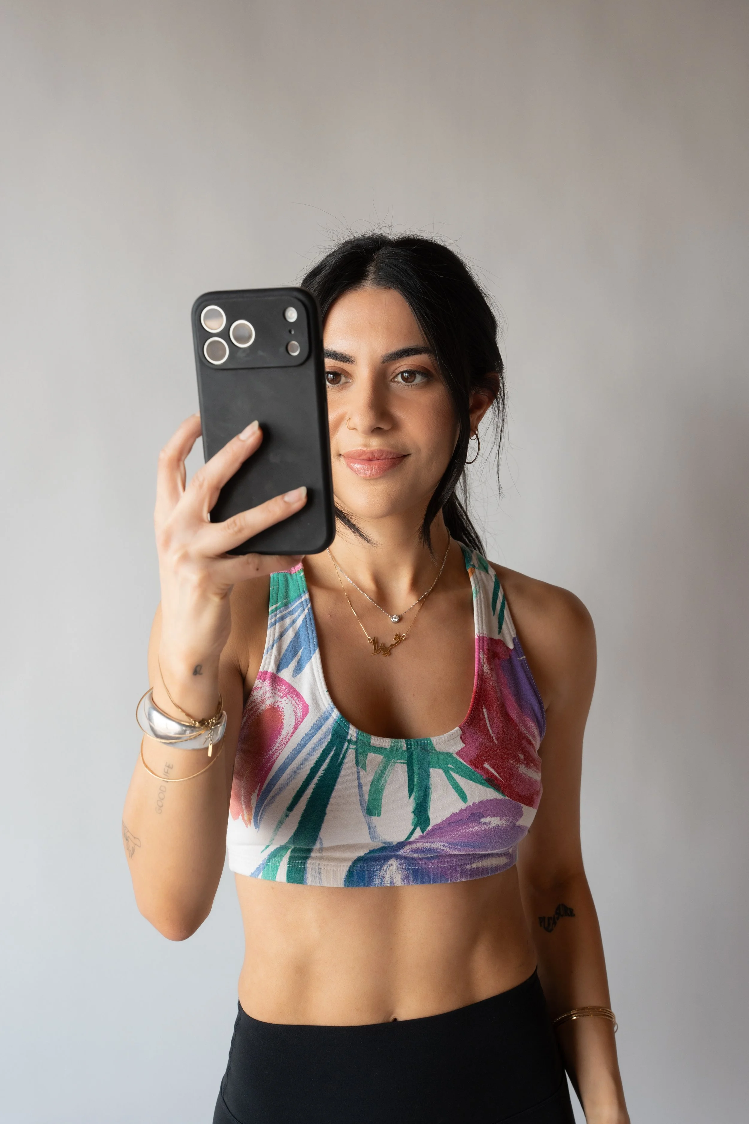 A woman with black hair wearing a colorful sports bra and black leggings taking a selfie with her smartphone against a plain background.