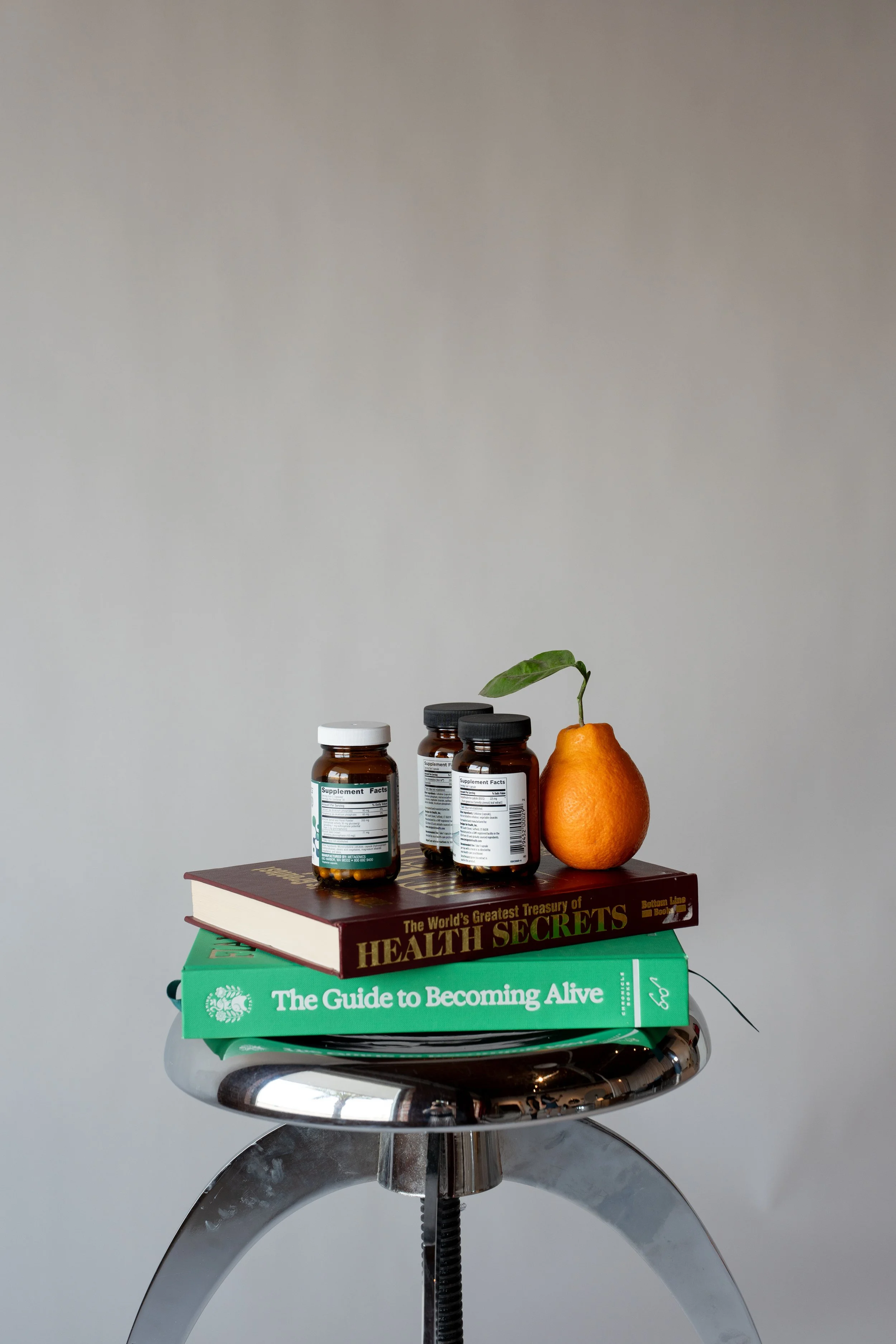 A stool holding three supplement bottles, a book titled 'The World's Greatest Treasure of Health Secrets,' a green book called 'The Guide to Becoming Alive,' and a pear with a green leaf.
