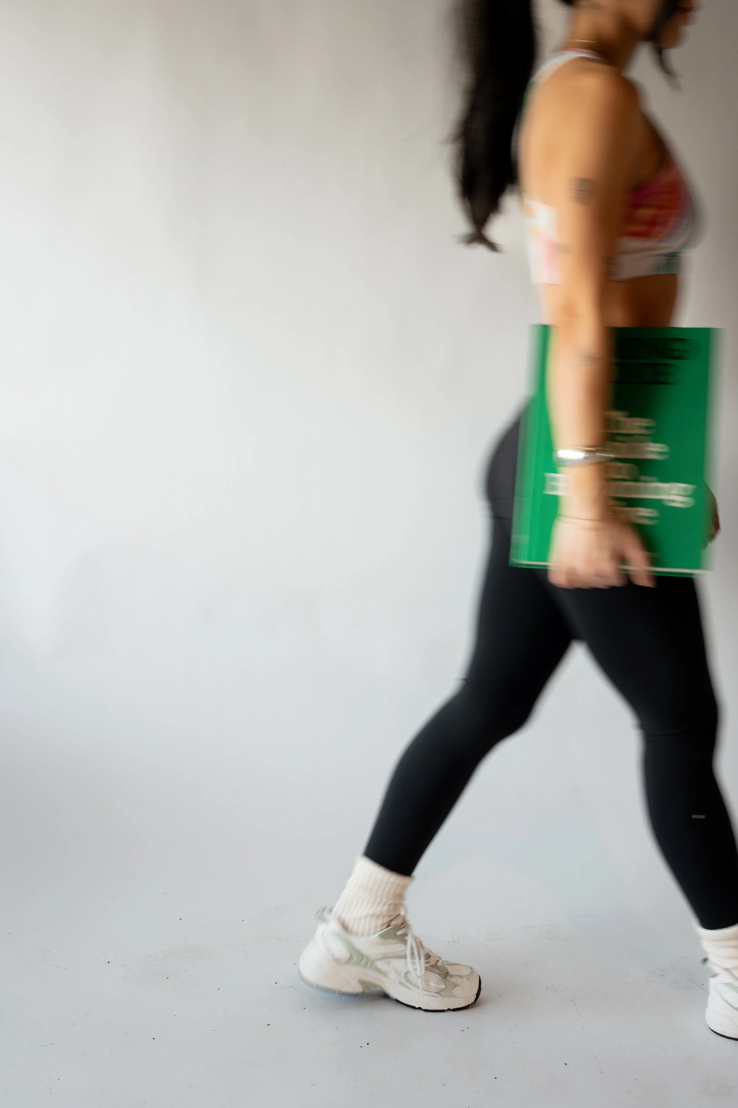A woman walking from the side, holding a green folder, wearing black leggings, white sneakers, and a colorful sleeveless top, with messy dark hair and a tattoo on her upper arm.