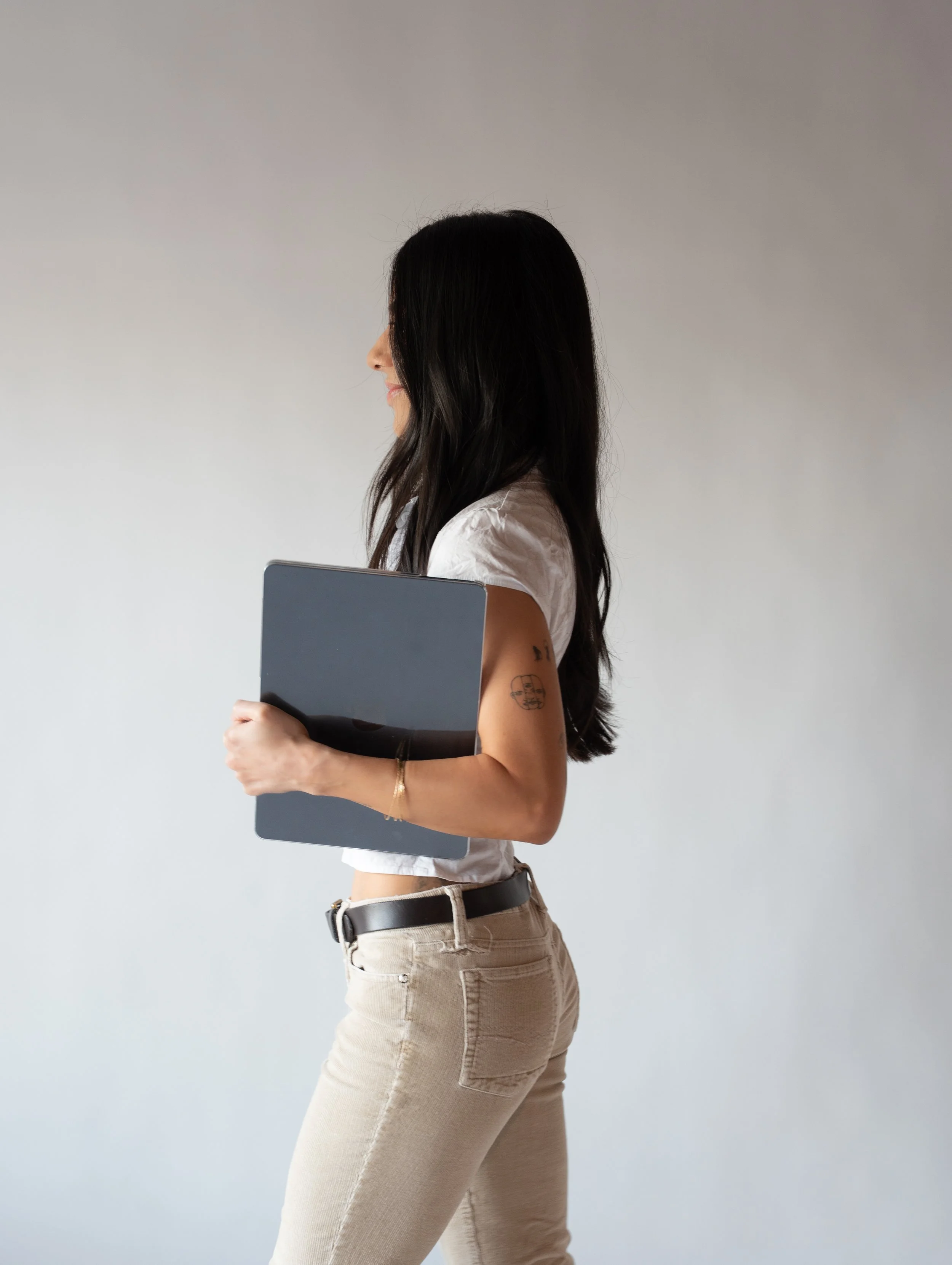 Side view of a woman with long black hair holding a closed laptop against her chest, wearing a white top and beige pants with a black belt, standing against a plain white background.