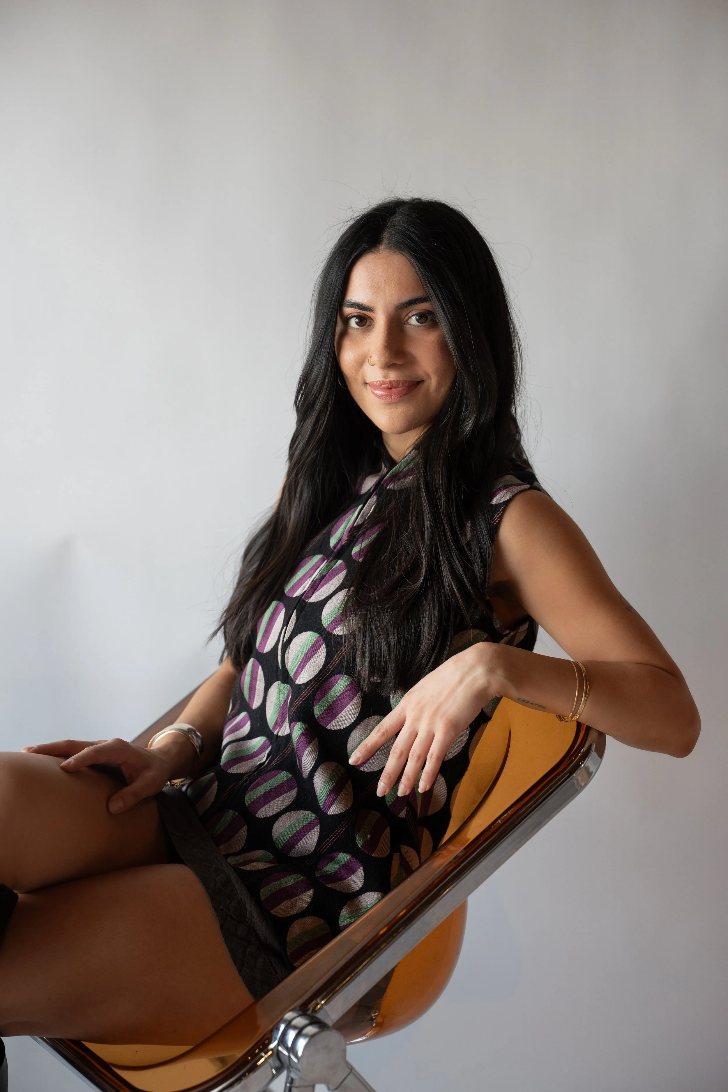 A woman with long black hair and a nose piercing, sitting on an orange chair, wearing a patterned sleeveless top, smiling at the camera.