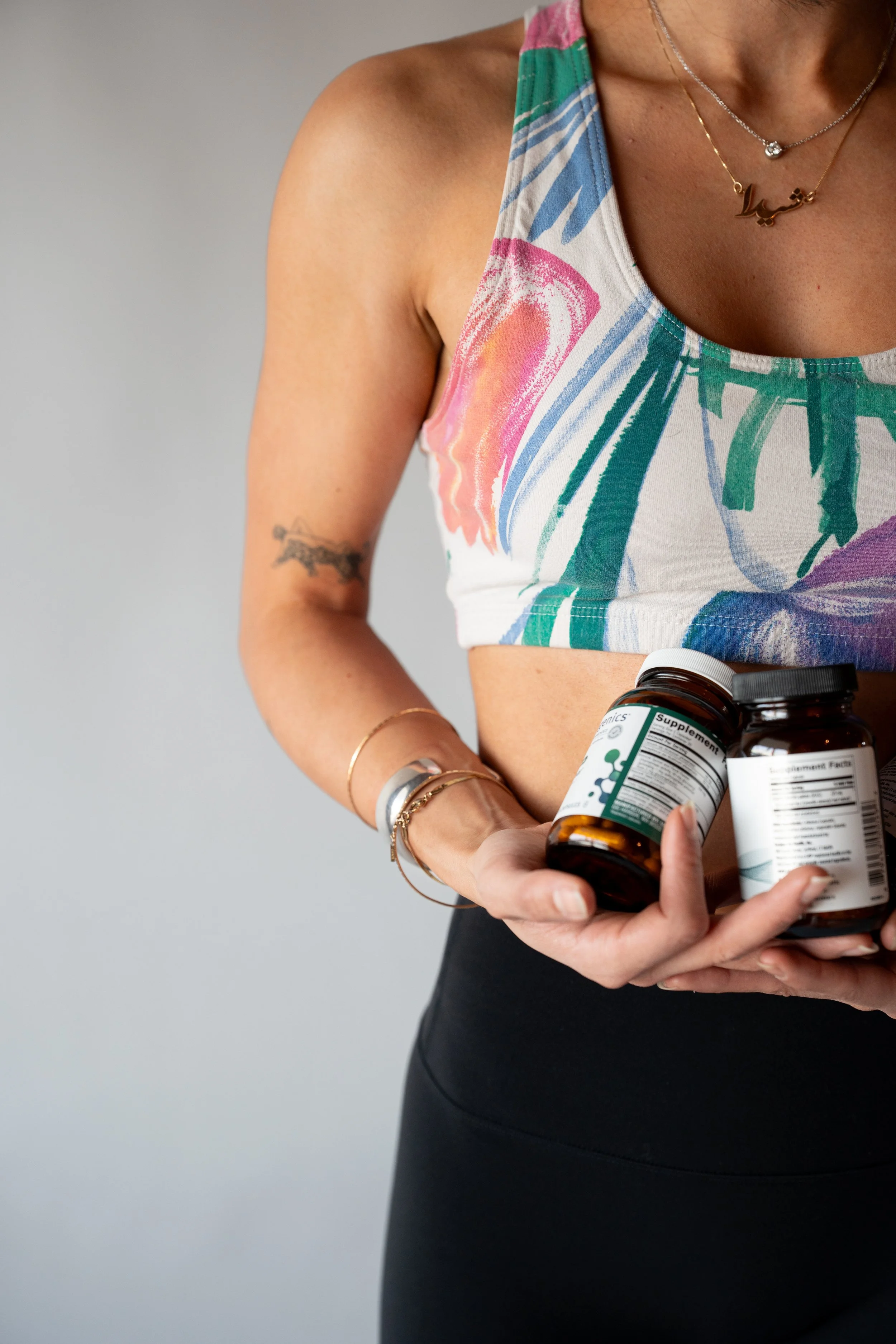 A woman holds two bottles of dietary supplements, wearing a colorful sports bra and black leggings, with jewelry on her arms and neck.