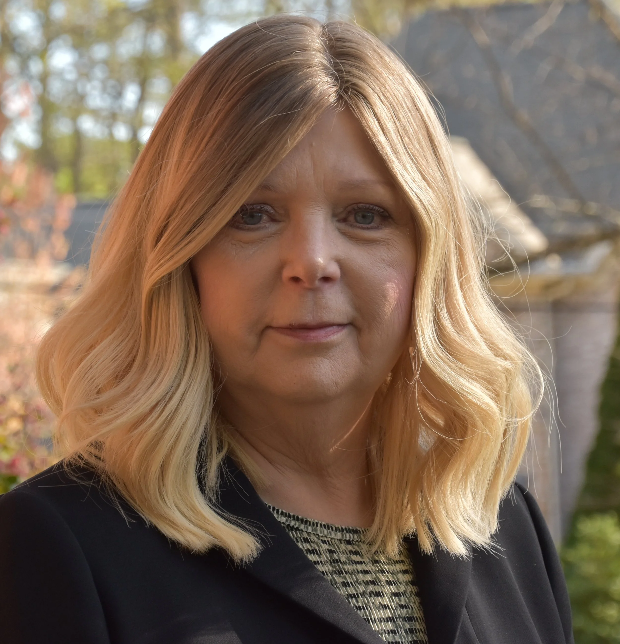 Close-up of a woman with blonde hair, wearing a black top with a decorative collar, outdoors in a natural setting with blurred trees in the background.