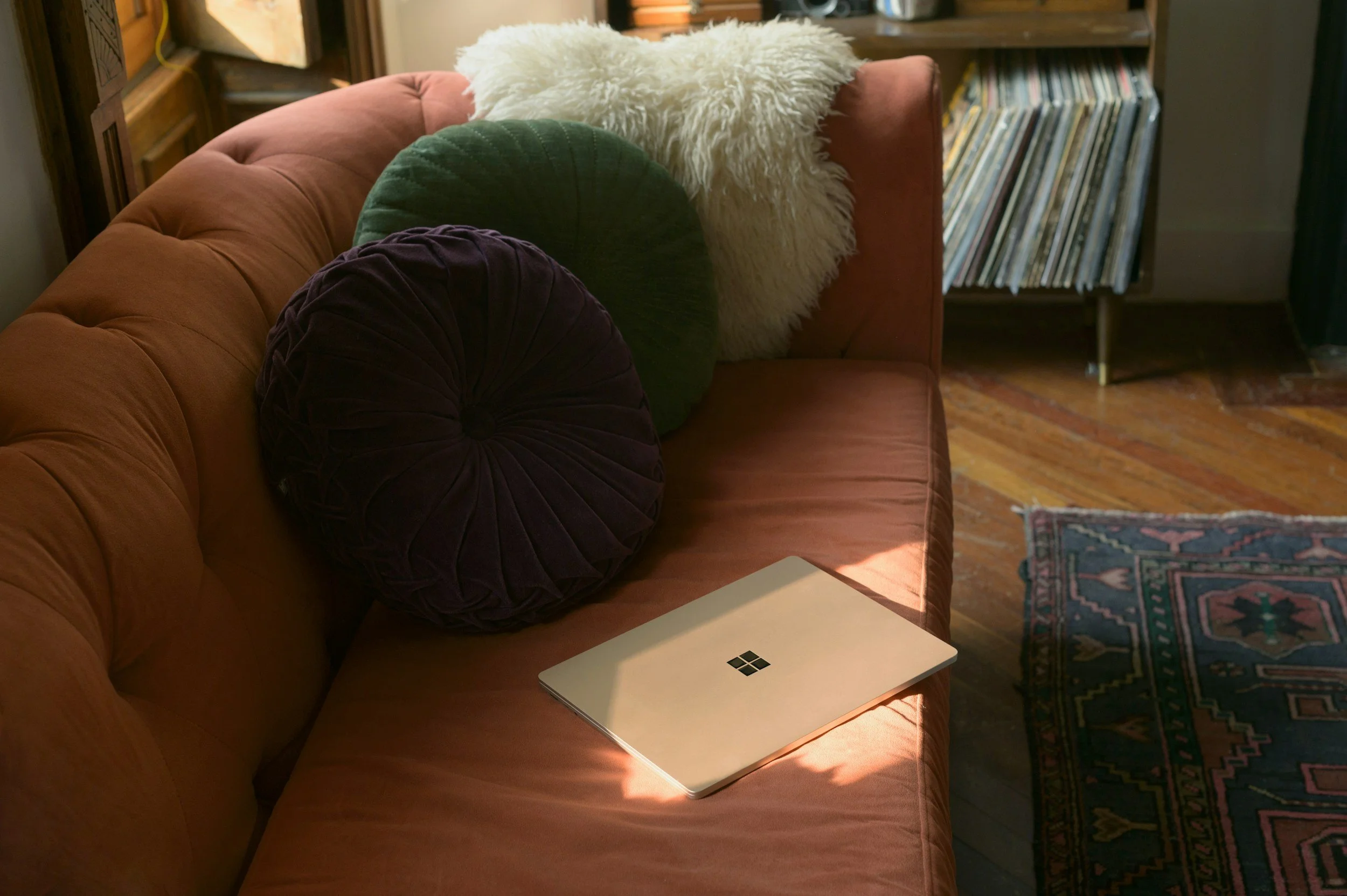 A pink couch with two decorative pillows, one white fluffy, one dark green, and one round dark purple. A closed laptop with a Windows logo is on the couch. To the right, a bookshelf with vinyl records, and a colorful rug on a wooden floor.