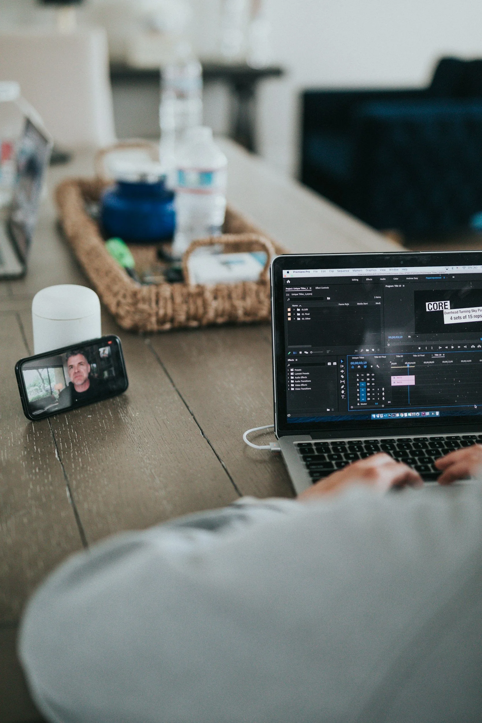 A person working on a laptop editing video with audio editing software open, a smartphone with a man’s face on the screen recording or participating in a video call, and various items on a wooden table including water bottles, a basket, and a white s