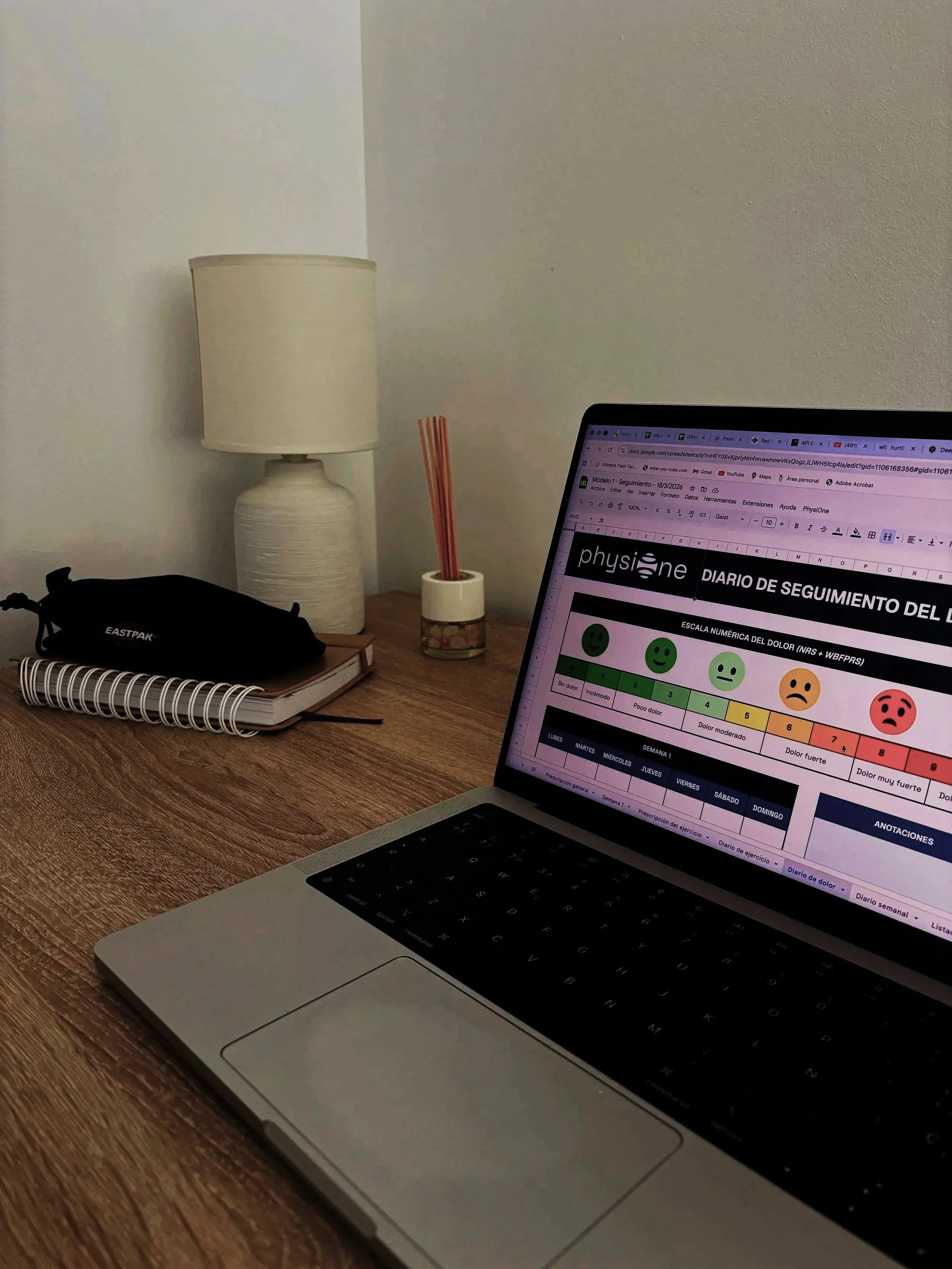 Desk with a closed notebook, black pencil case, and spiral notebook, a desk lamp, and a small container with orange sticks, with a laptop open displaying a health tracking spreadsheet in Spanish.