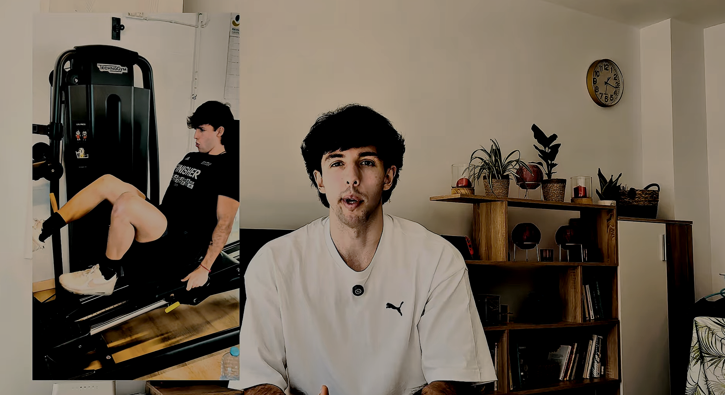 A man with dark hair wearing a white Puma shirt sitting in a living room. Behind him, a wooden shelf with potted plants, books, and decorative items. A clock on the wall shows around 12:12. To the left, a large poster of a young man exercising on gym