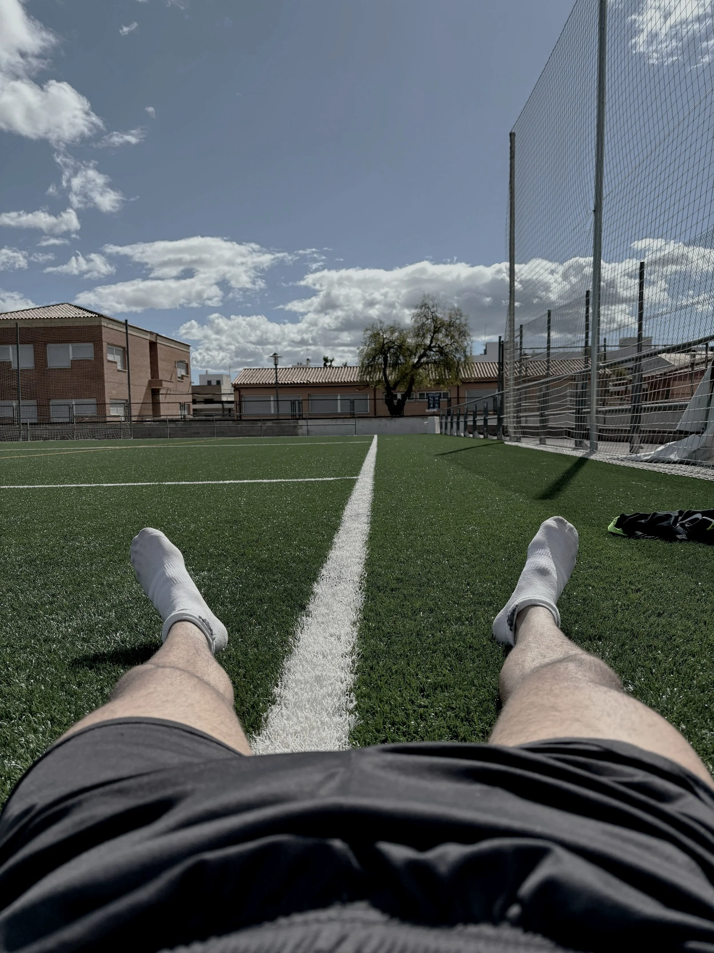 Person lying on a football field, legs extended, view of the field, goal post, and buildings in the background under a partly cloudy sky.