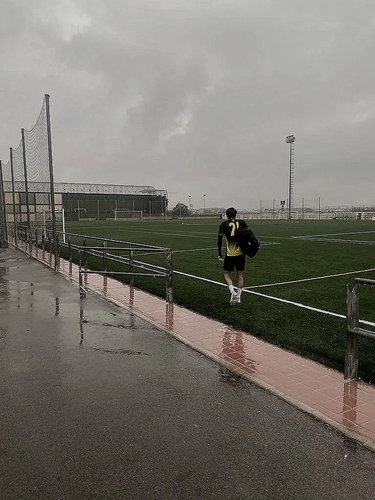 A person walking along a wet track beside a soccer field on a cloudy day, carrying a backpack and wearing sports attire.
