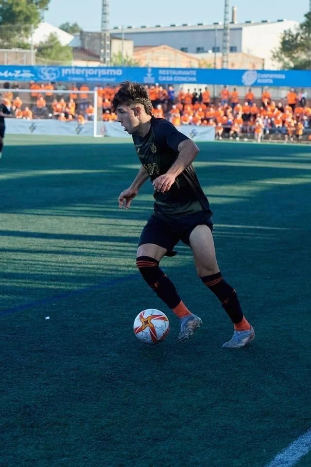 A young male soccer player in black uniform with orange and black socks is on a green soccer field, controlling a white and red soccer ball, with a crowd of spectators in the background.