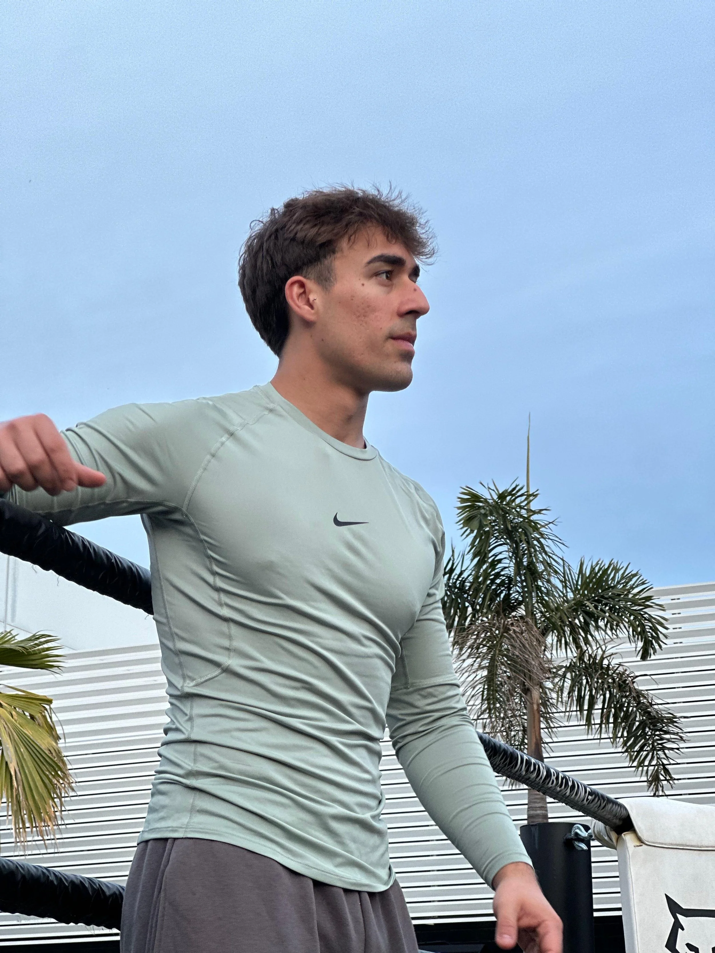 A young man with brown hair and an earring in his left ear stands outdoors near palm trees and a modern building with horizontal lines, wearing a gray Nike long-sleeve shirt and gray pants.
