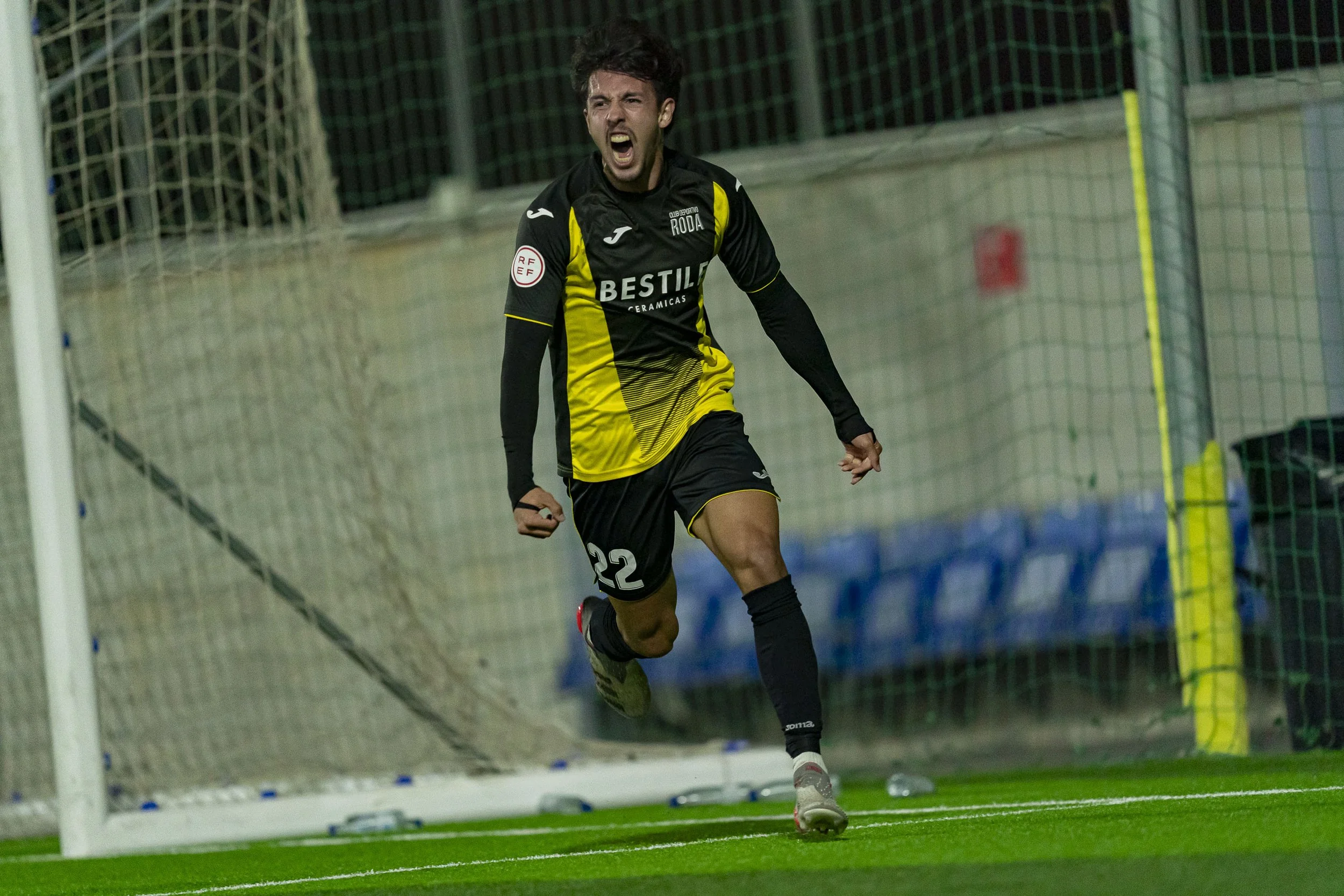 A soccer player celebrates a goal on the field, wearing a black and yellow uniform with the number 22, near the goalpost.