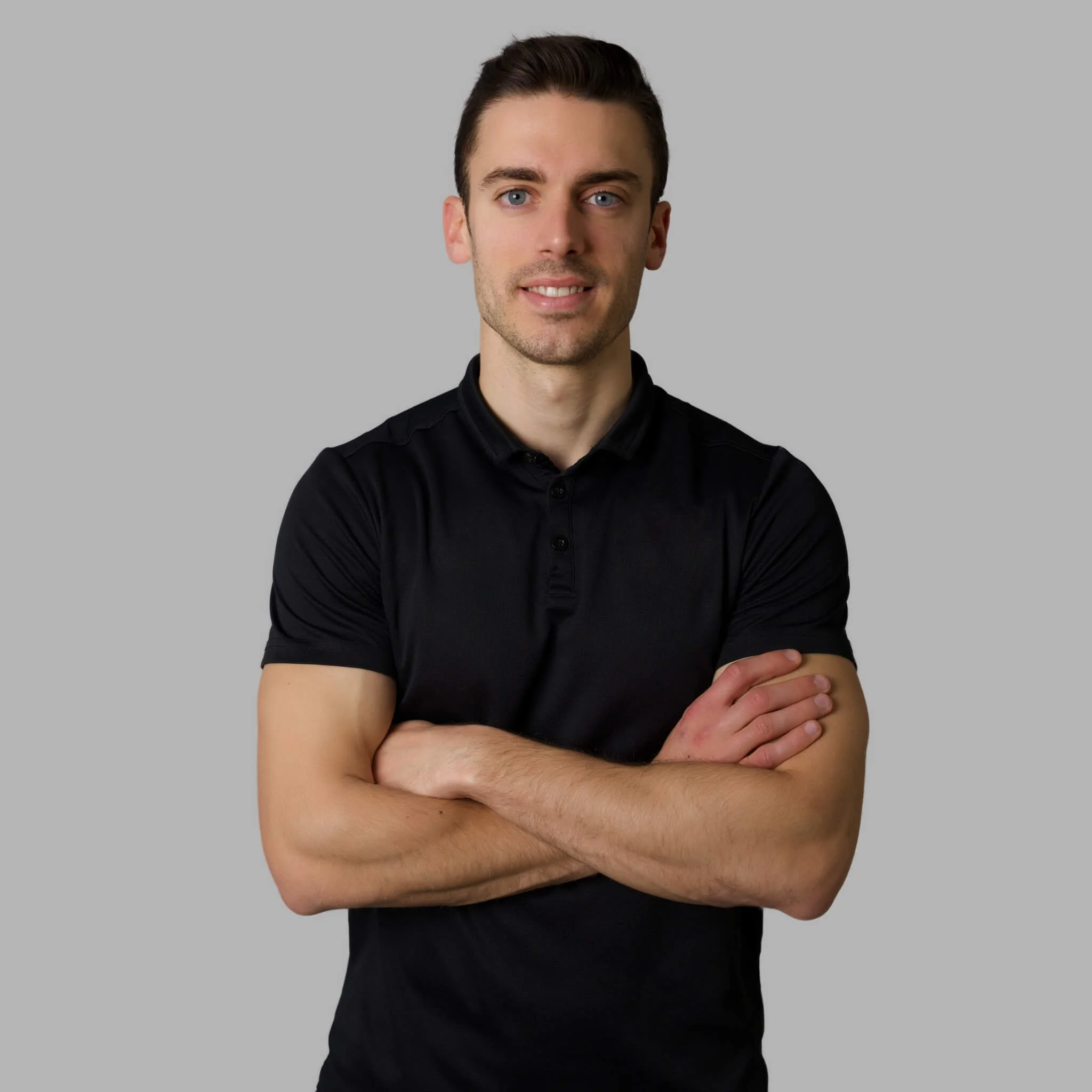 A young man with dark hair, blue eyes, and light skin, wearing a black short-sleeved polo shirt, crossing his arms and smiling at the camera against a plain gray background.