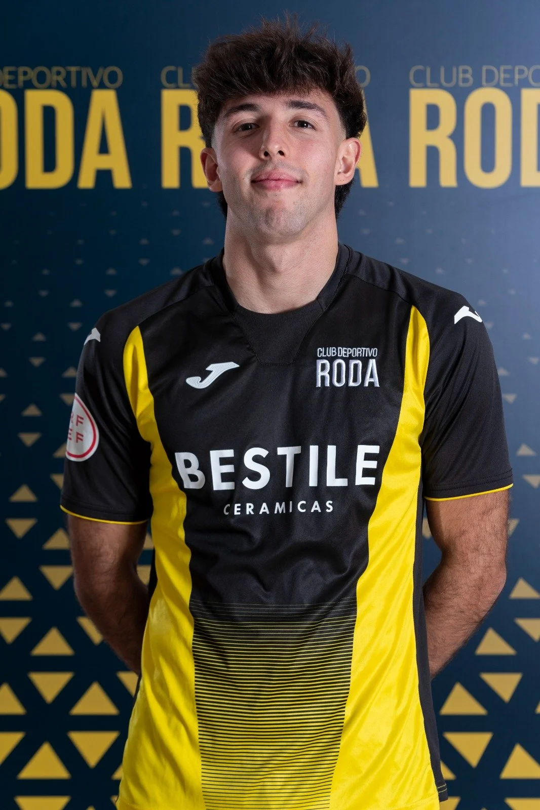 Young man in black and yellow sports jersey standing in front of a blue background with yellow text, smiling at the camera.