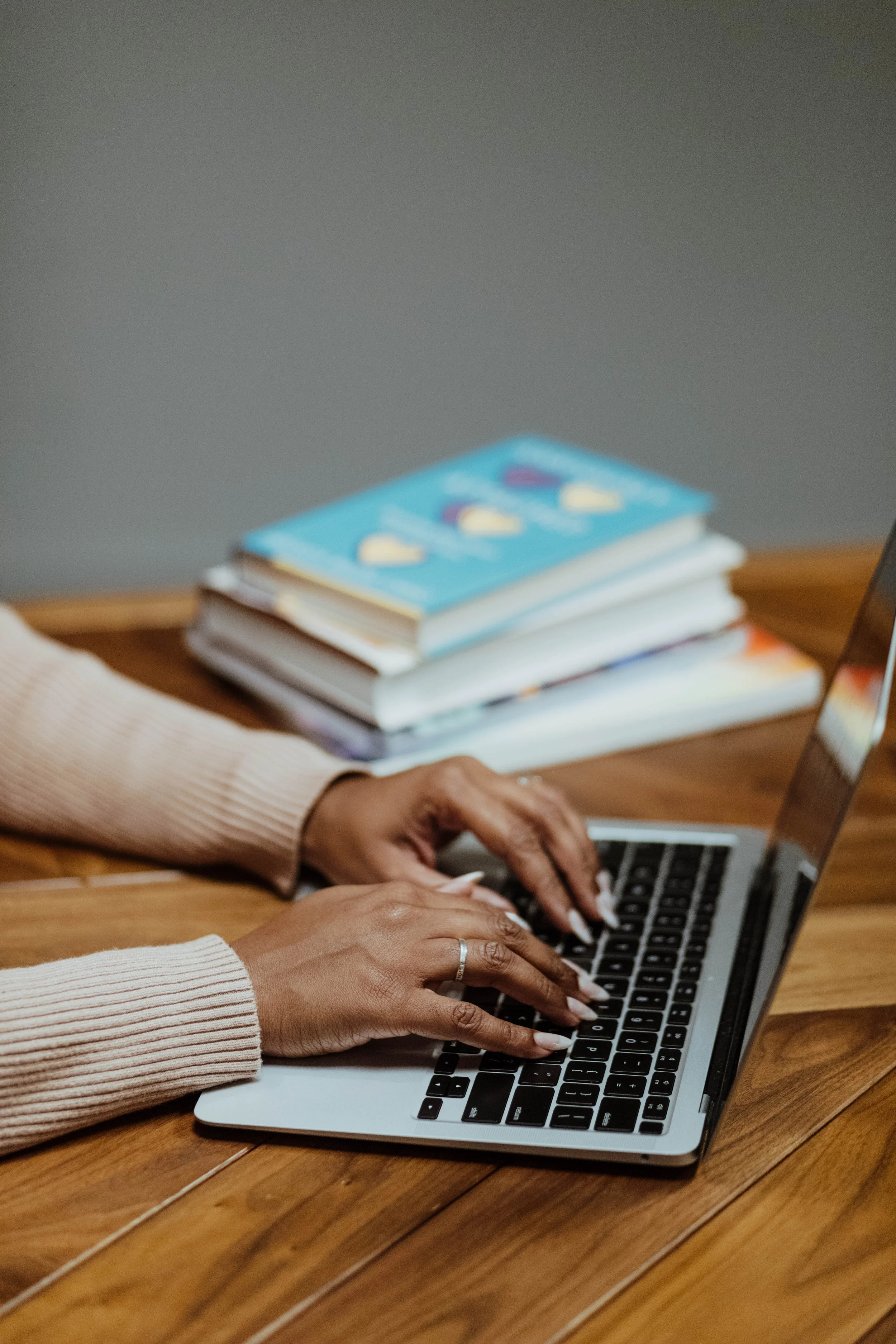 Close-up of a person typing on a laptop keyboard, with a stack of books on a wooden table nearby.
