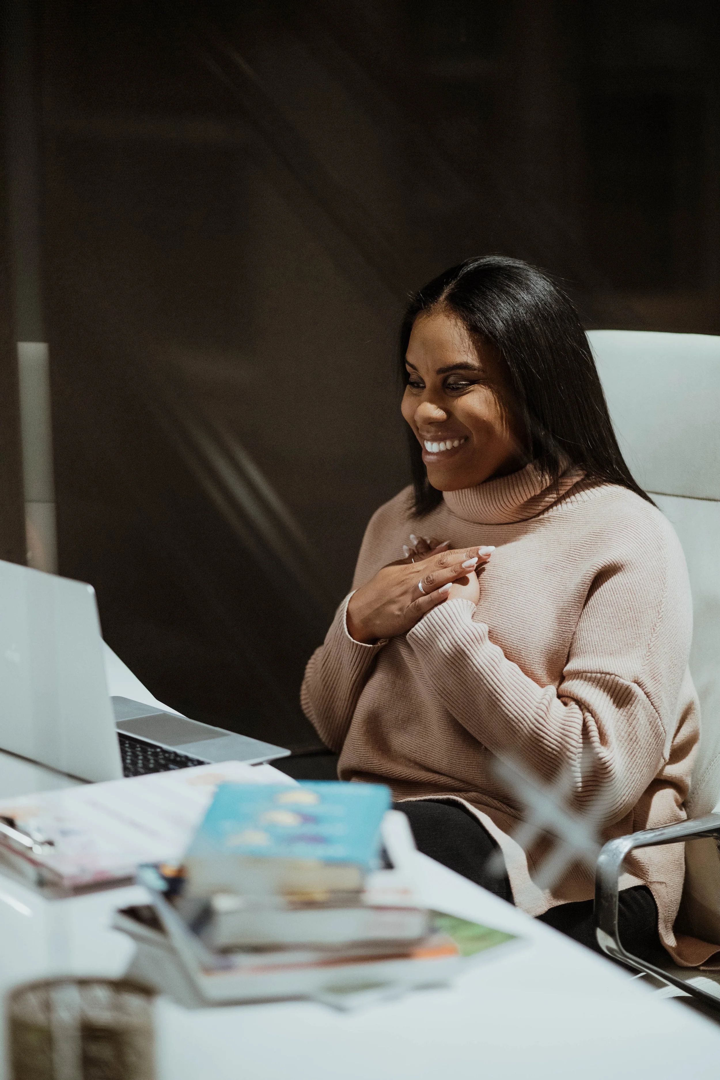 Smiling woman sitting at a desk with a computer, hands clasped over her chest, in a modern office setting.