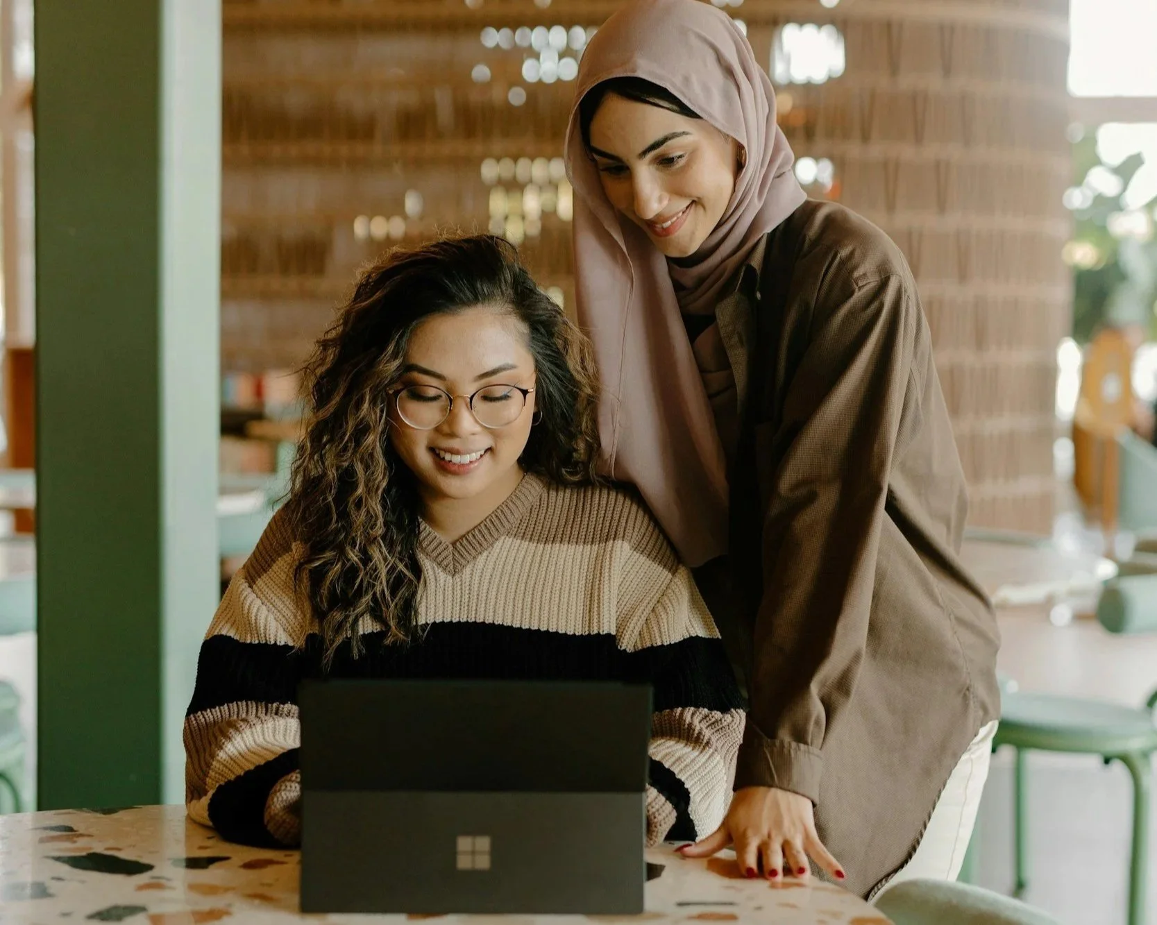 Two women working together at a laptop in a cozy, well-lit cafe. One woman with curly hair and glasses is seated, while the other woman in a hijab leans over showing her something on the laptop.