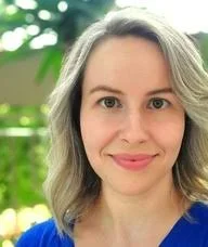 Psychotherapy student Joanna Sulzycki. White woman in her 30s with shoulder-length blonde wavy hair and hazel eyes; she smiles at the camera. She has on a royal blue v-neck cotton shirt and is outdoors with sunshine and greenery (blurred) behind her.