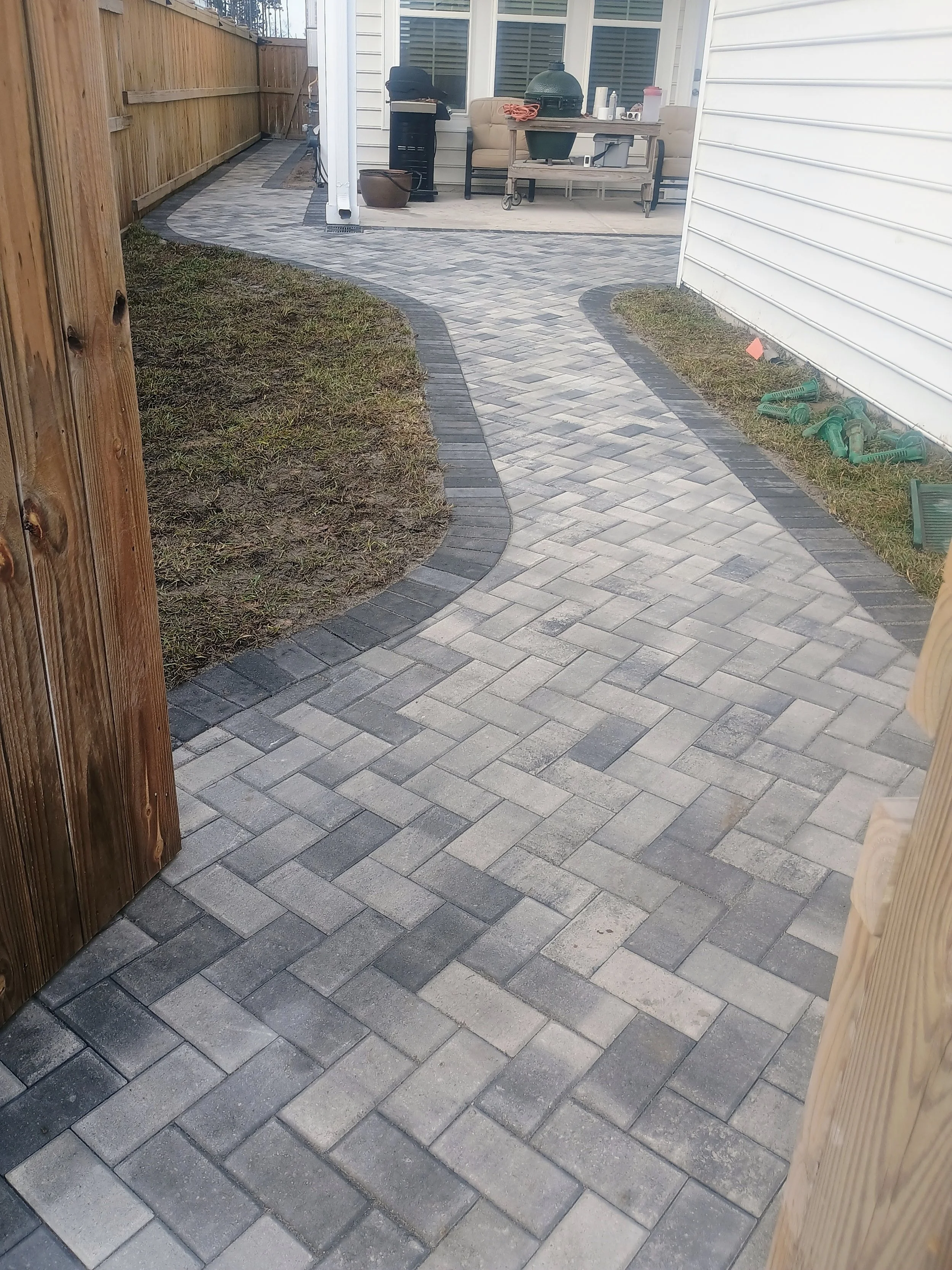 Newly paved outdoor walkway with interlocking brick pattern, bordered by dark bricks, leading from a fenced yard to a patio area with seating and barbecue grill.