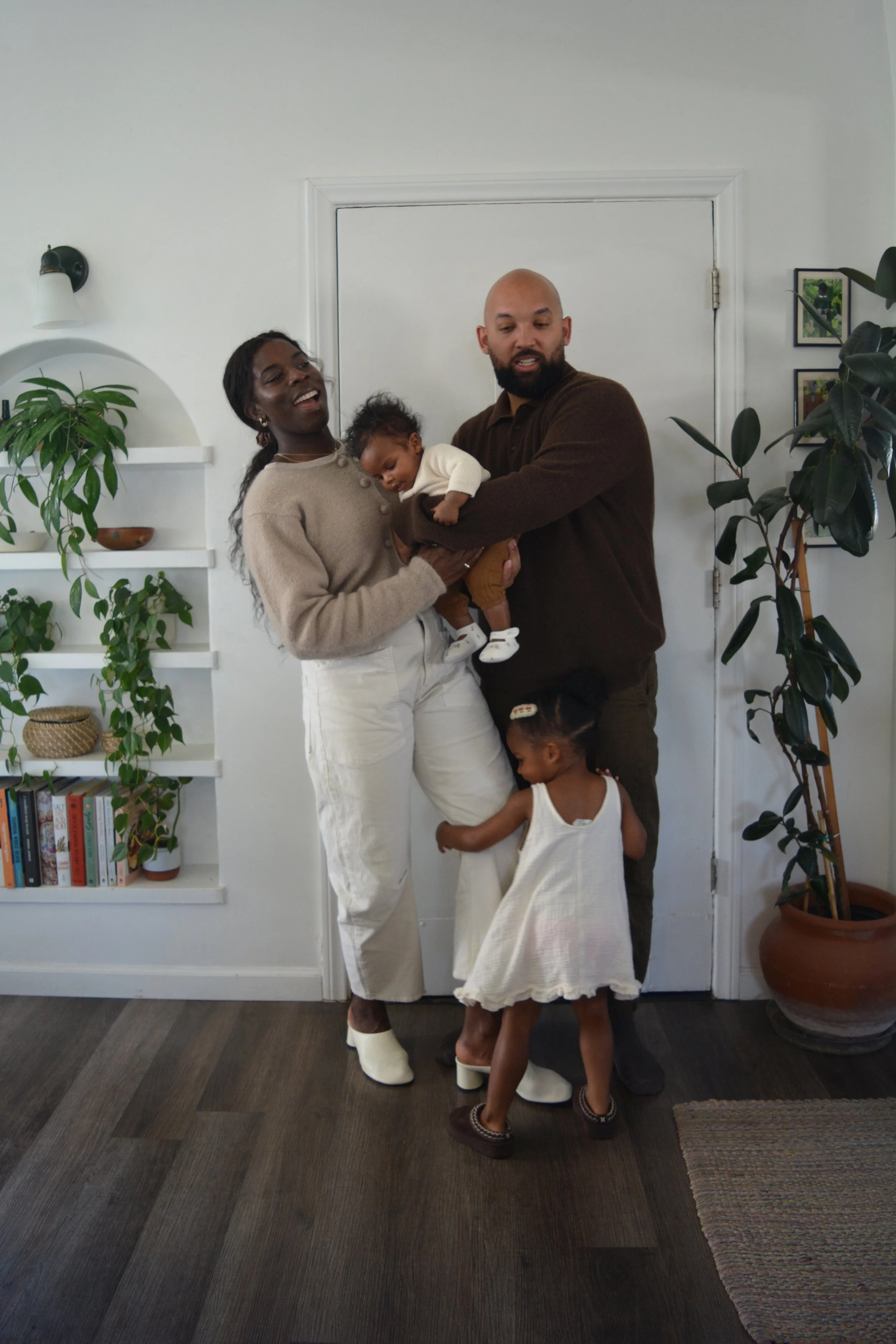 A Black woman with long hair, white pants, and a beige sweater holding a baby and smiling, standing next to a light-skinned man with a beard, brown sweater, and white shoes, who is holding a young girl in a white dress. They are in a room with wall-m