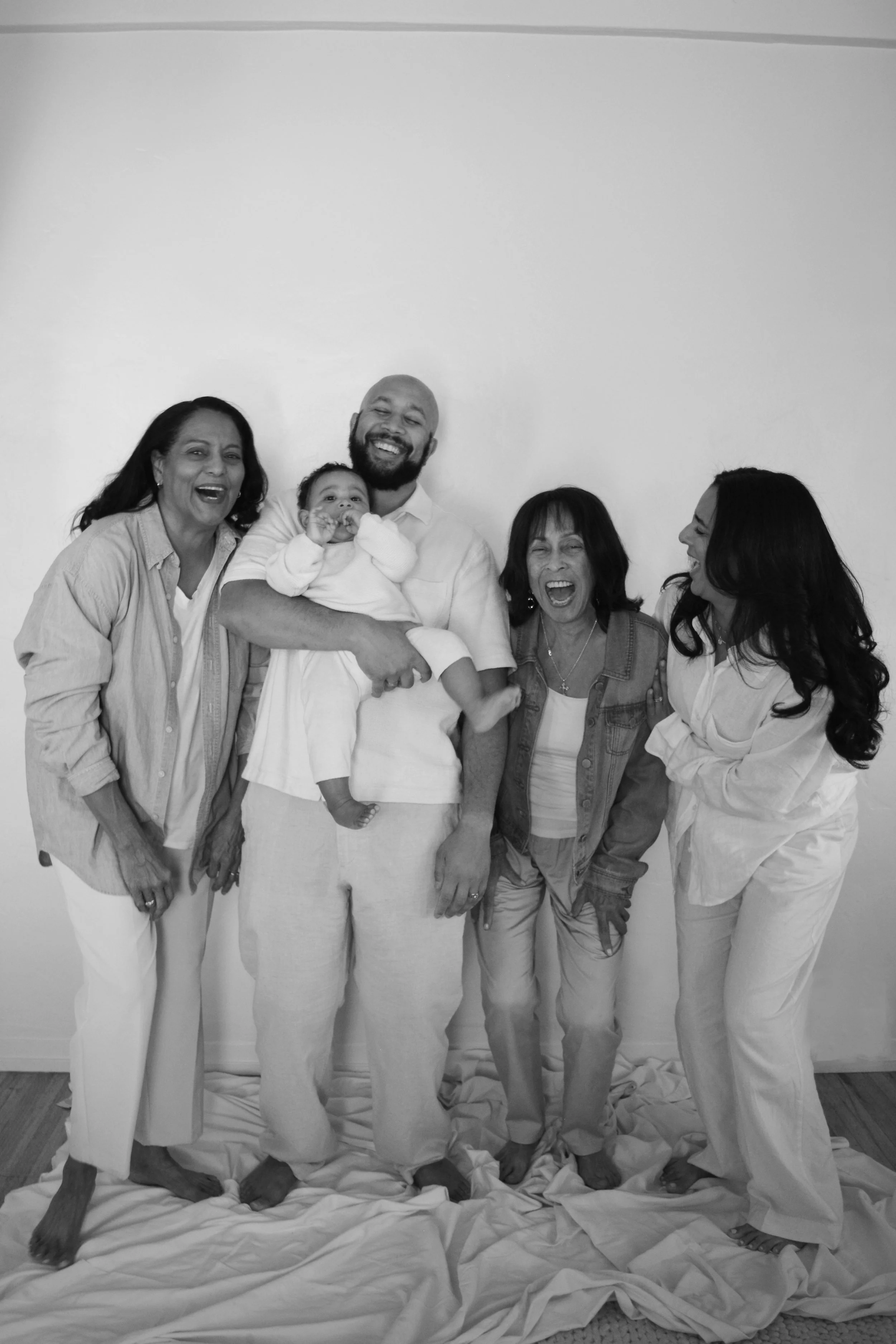 Group of six people laughing and smiling, standing on a sheet on the floor against a plain wall, with a baby being held in the center.