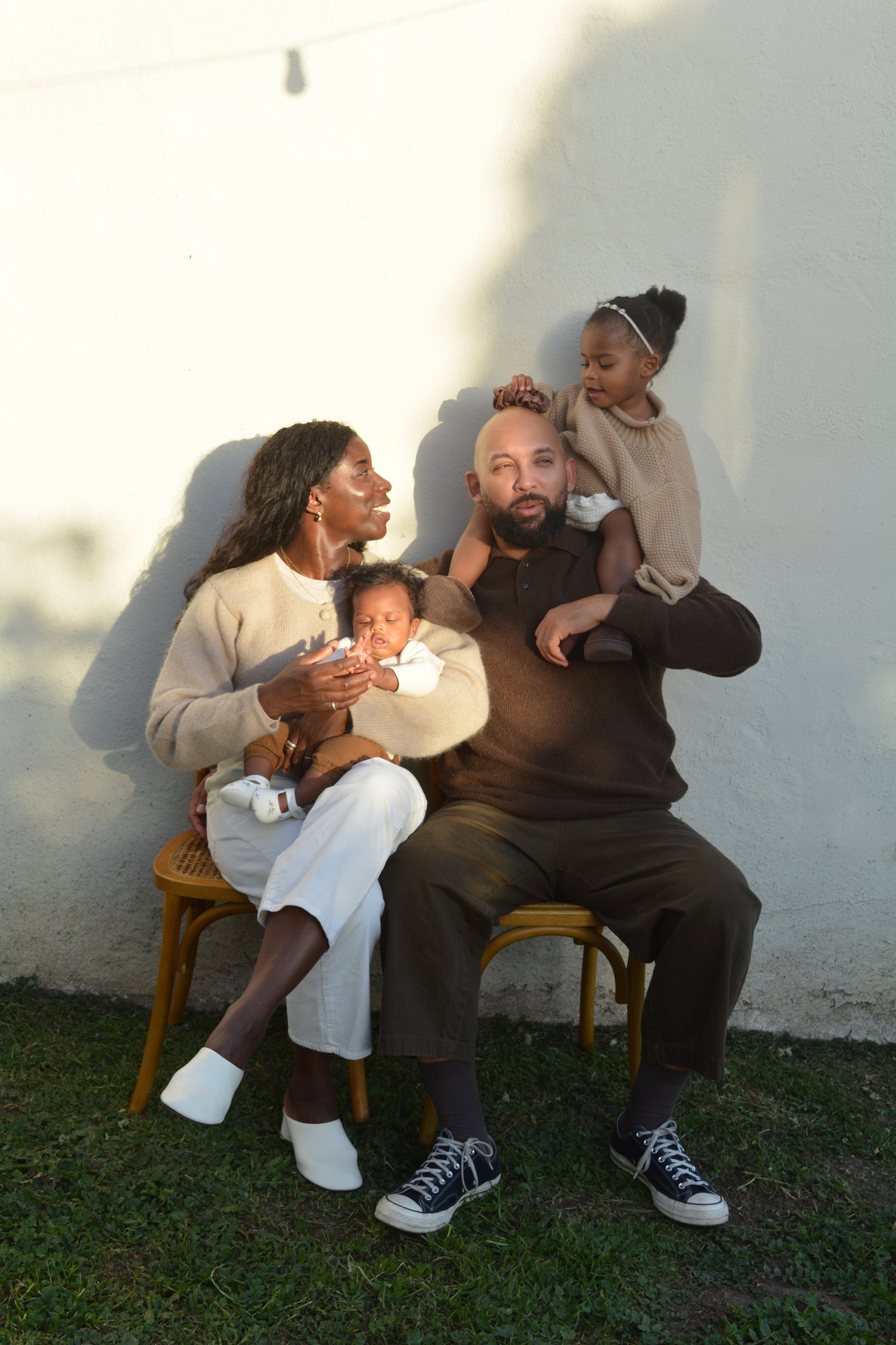 A family of four sitting outdoors against a plain wall. The woman, holding a baby, is smiling at a man who is carrying a young girl on his shoulders. The man is wearing black sneakers, and the woman is in white shoes. The girl has her hair styled in 