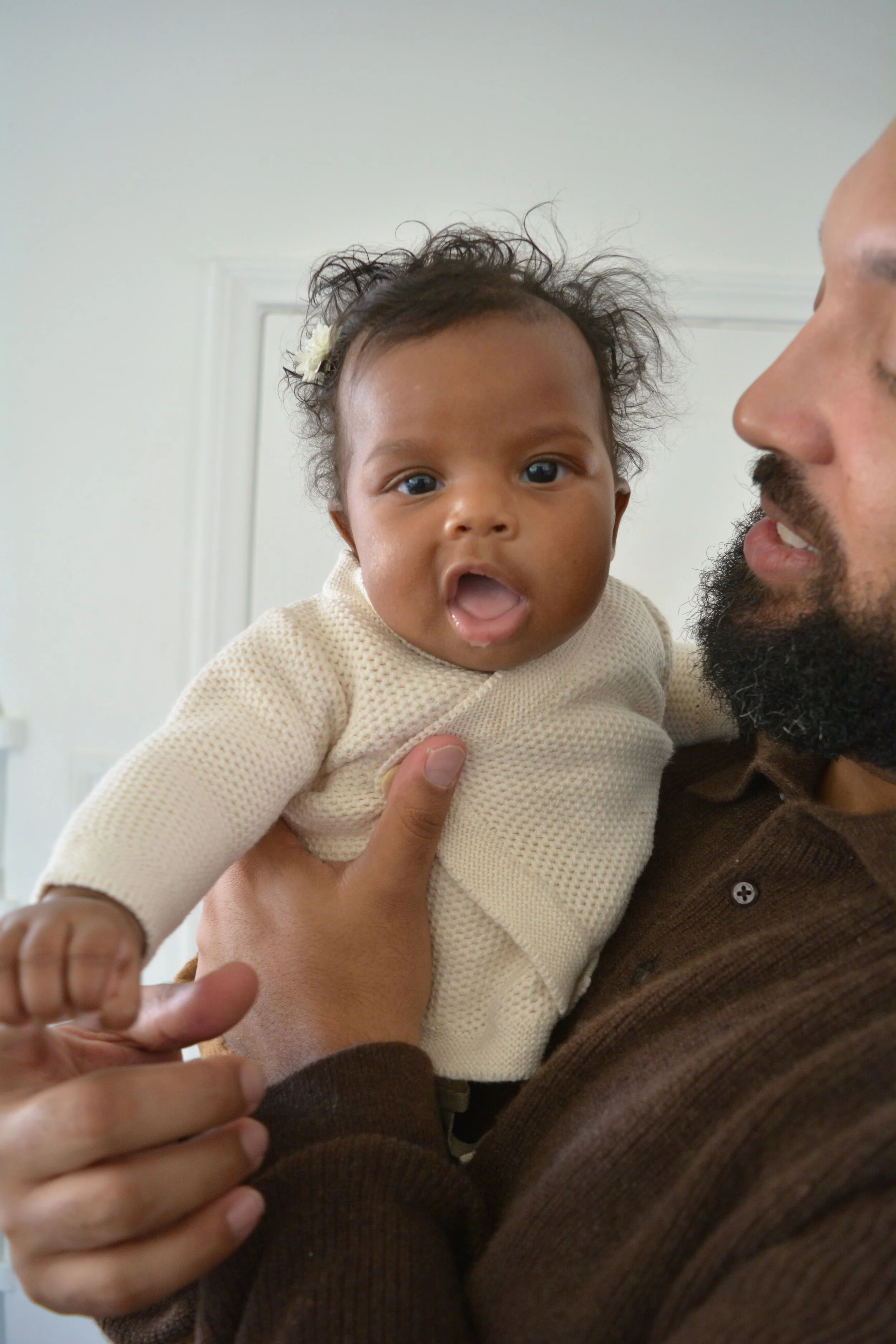 A man holding a young girl with curly hair and a flower clip, close to his face, indoors with beige walls.