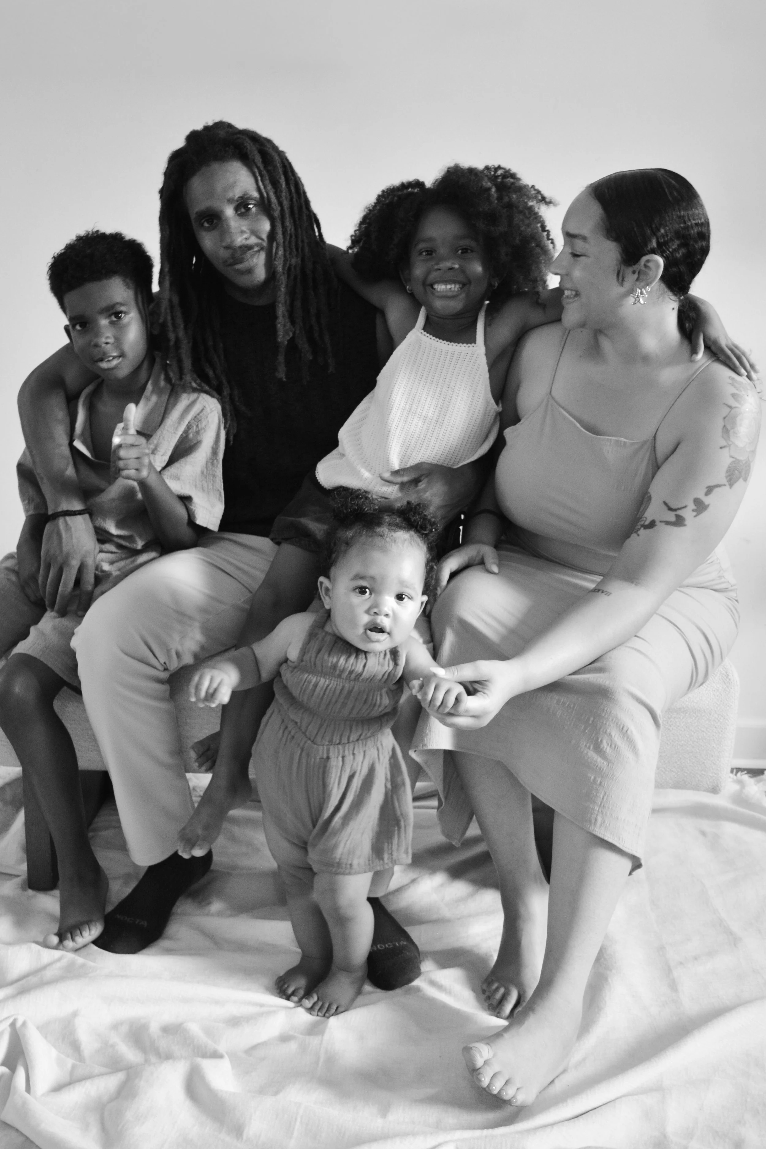 A diverse group of six people, including children and adults, sitting together on a couch. They are smiling and appear happy. The image is in black and white.