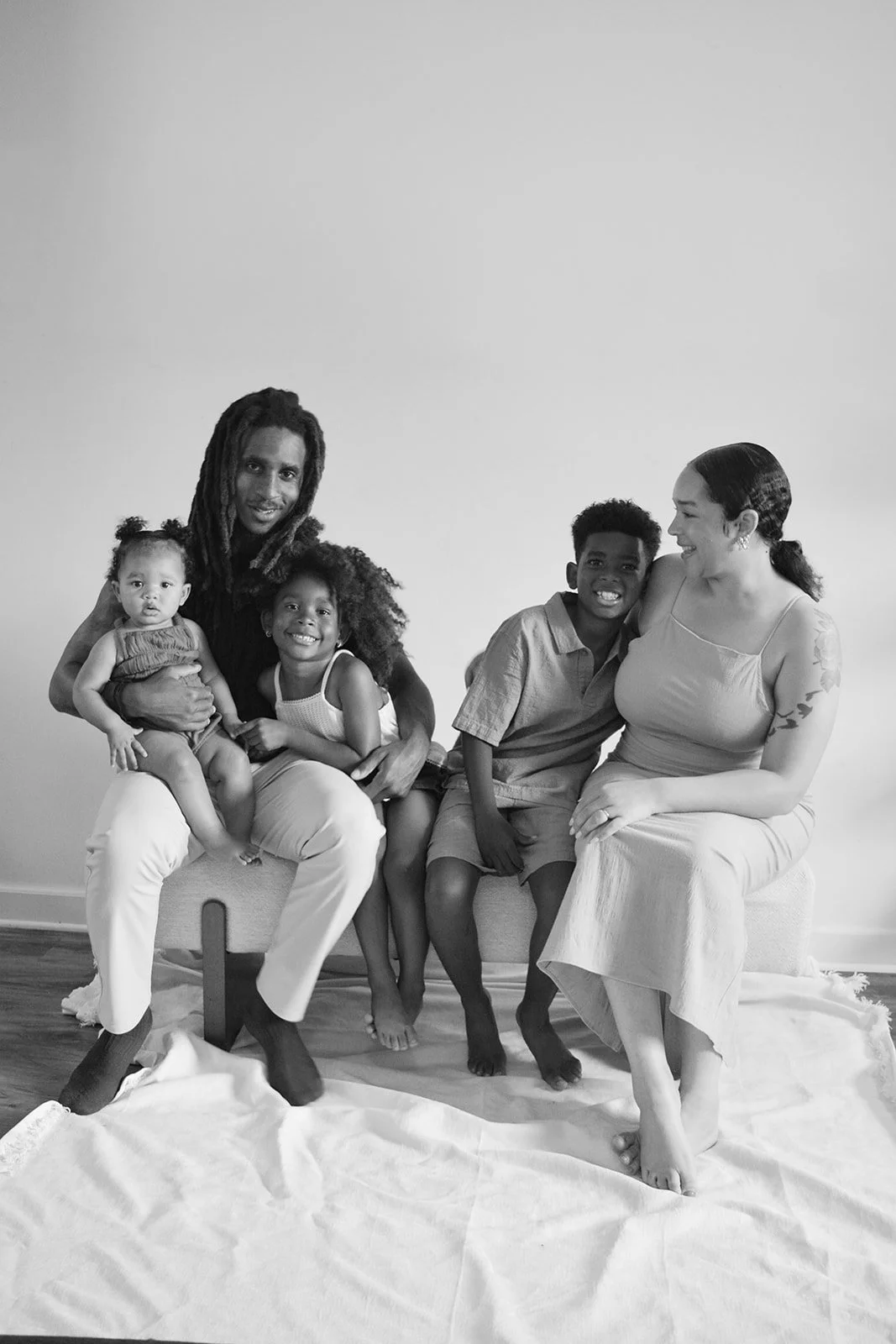Family portrait with six members, two adults and four children, sitting together on a light-colored blanket on the floor in front of a plain wall, smiling and interacting warmly.