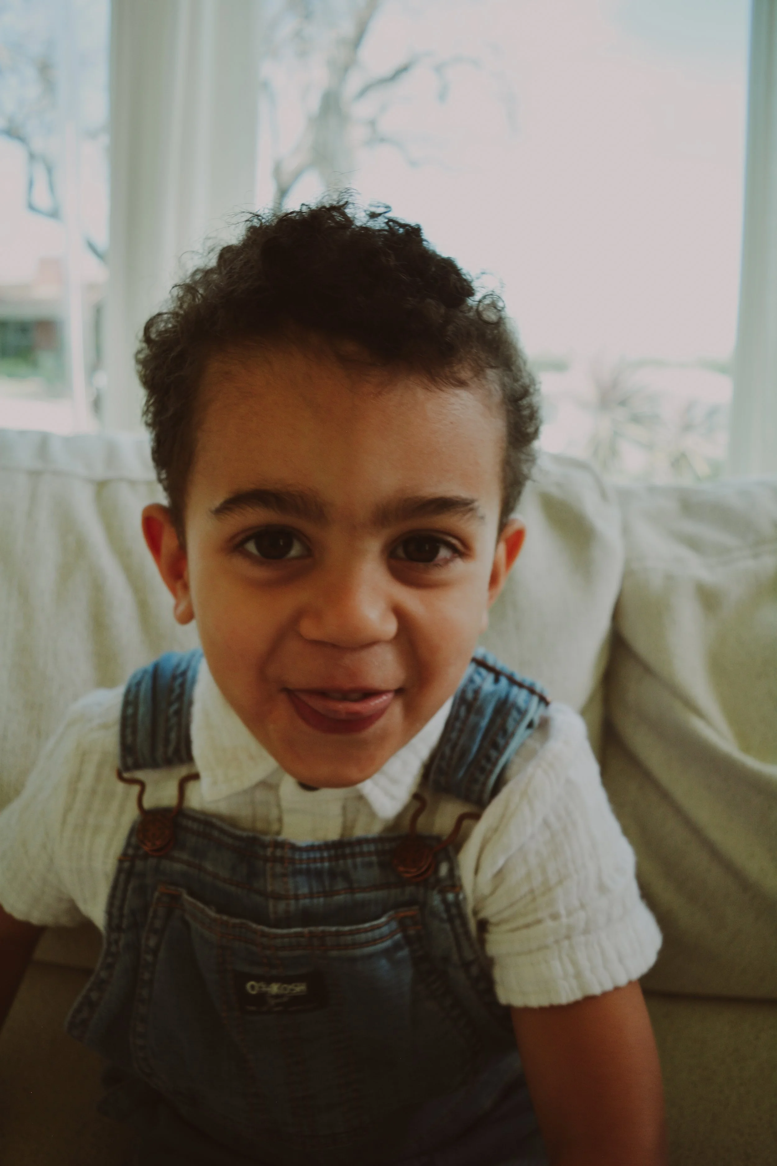 A young boy with curly hair and big eyes smiling, wearing a white shirt and denim overalls, sitting on a couch by a window with a tree outside.