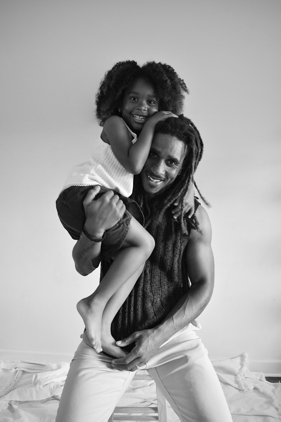 A man with dreadlocks holding a young girl on his shoulders, both smiling happily in a black and white photo.
