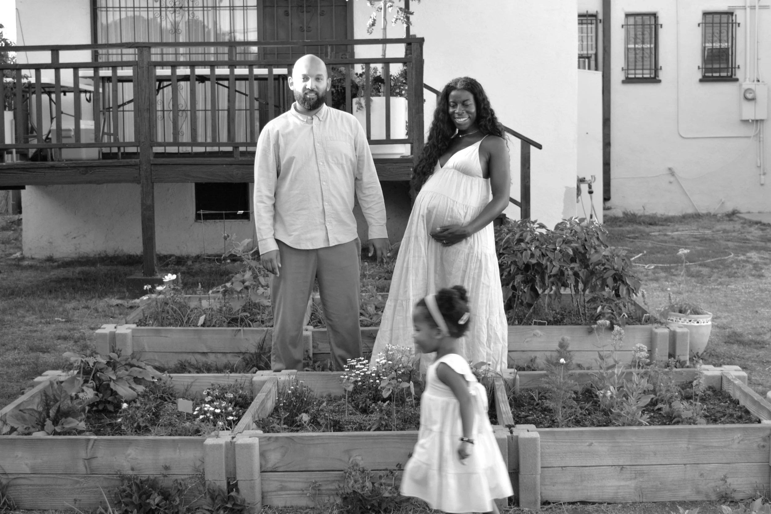 A man with a beard and a woman with long curly hair standing in a garden with raised flower beds, a house with barred windows in the background, and a young girl in a dress walking in the foreground.
