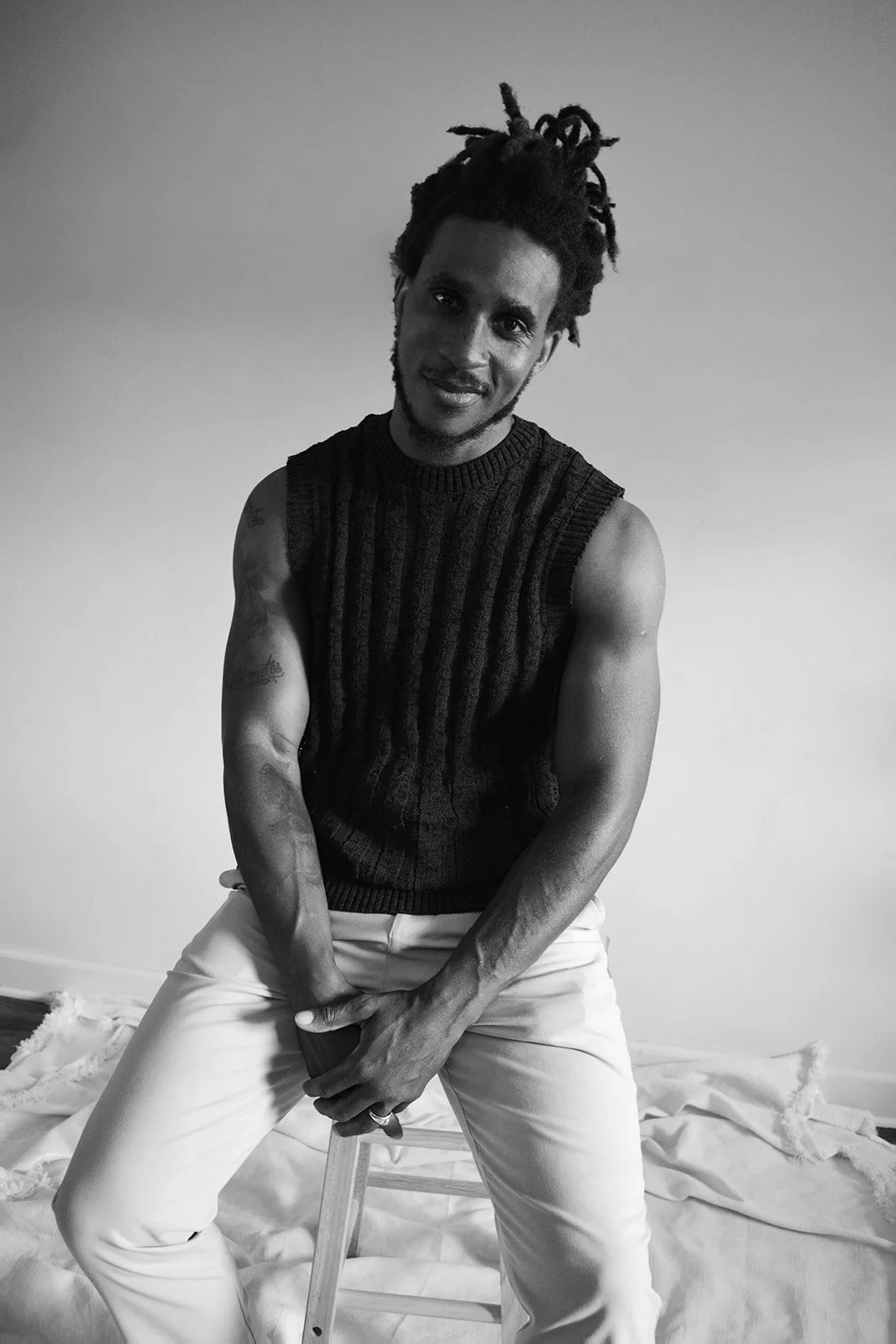 Black and white photo of a man with dreadlocks, wearing a sleeveless sweater, sitting on a stool, smiling at the camera, in a room with a plain wall and some fabric on the floor.