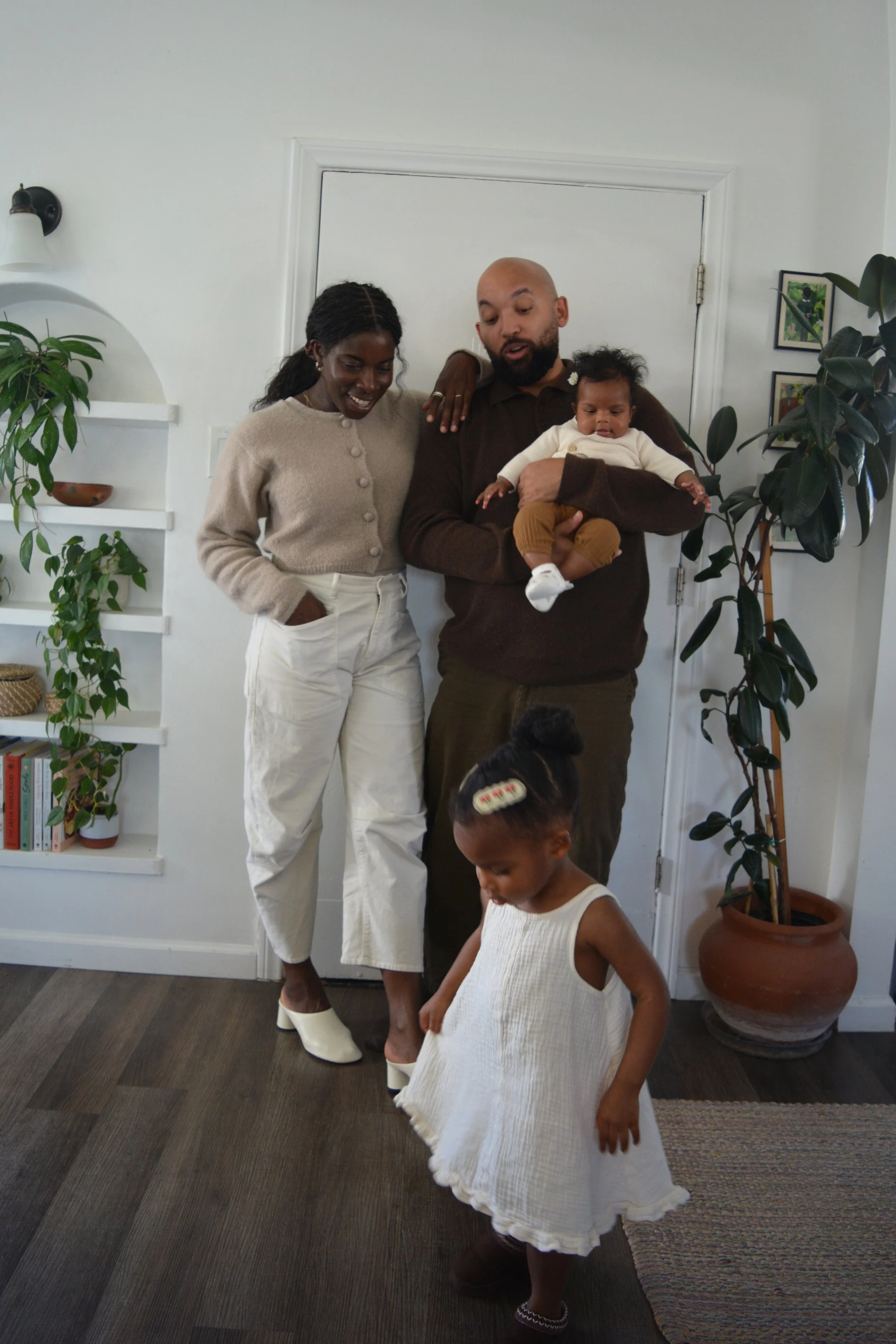 A family portrait featuring two adults and two children in a home setting. The woman is wearing a beige cardigan and white pants, standing with her hand in her pocket, smiling. The man is bald with a beard, wearing a brown sweater, holding a baby gir