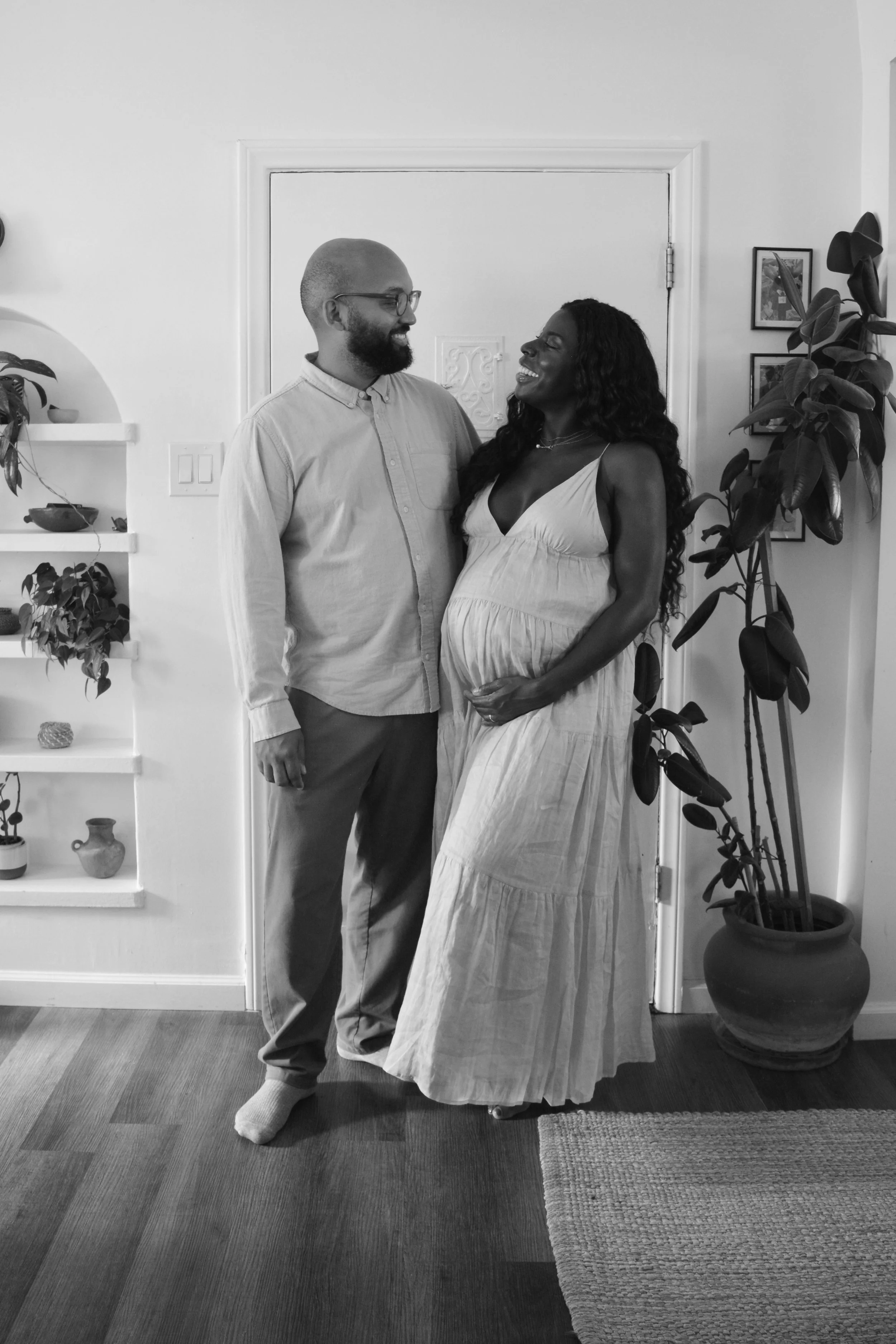 A happy couple, a man with glasses and a beard and a pregnant woman with long curly hair, smile at each other while standing in a home interior. The man is dressed in a light-colored shirt and trousers, and the woman in a long dress. They are in a co