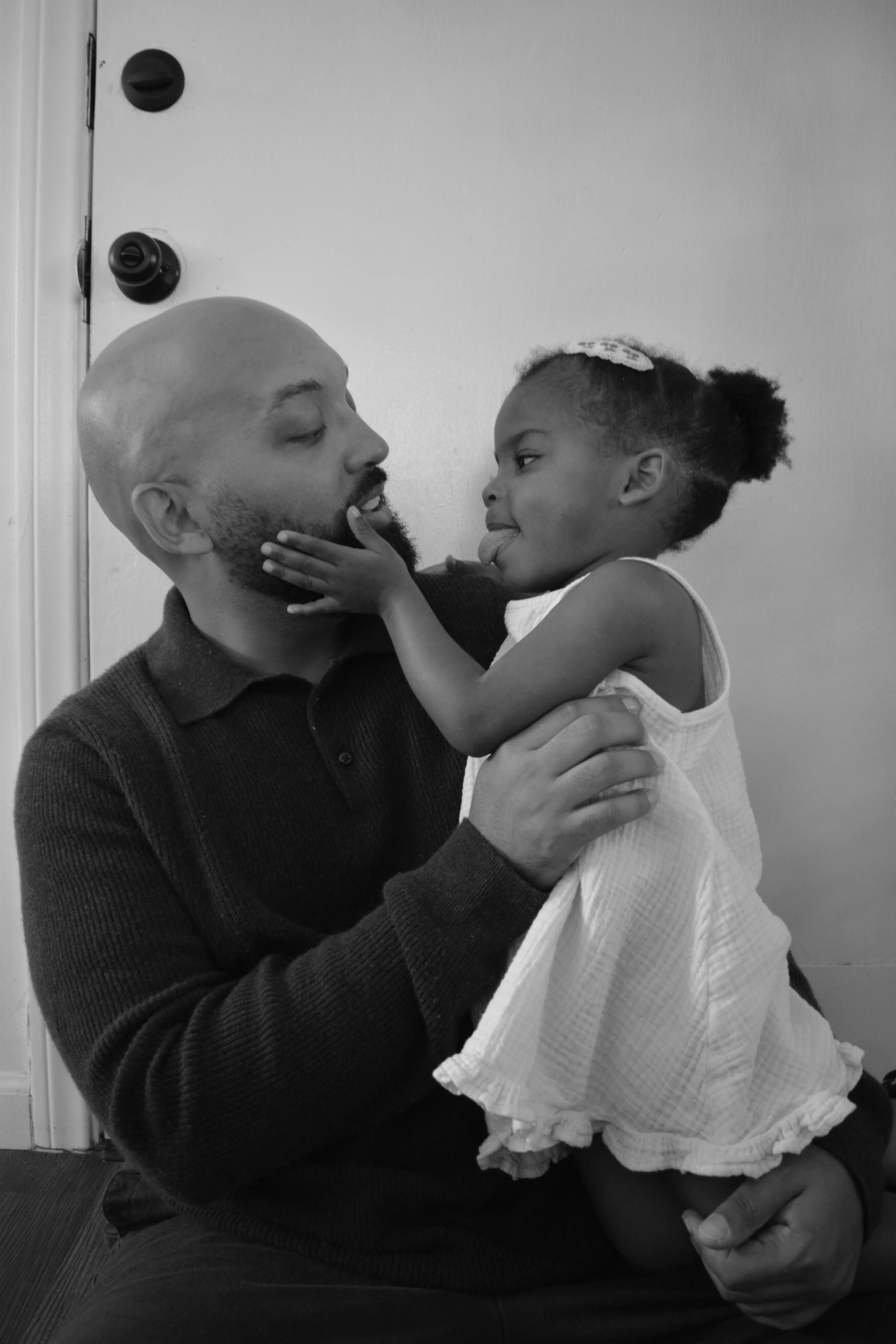 A man with a shaved head and a beard holding a young girl in a white dress. The girl has her tongue sticking out and is touching the man's face. The man has his eyes closed and a gentle expression.