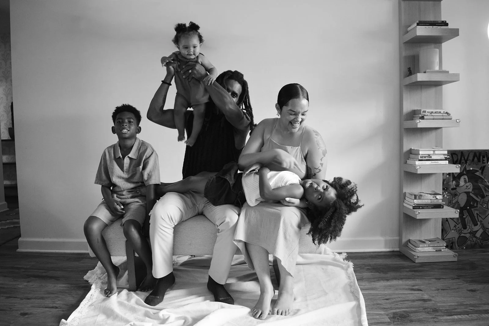 A joyful multi-ethnic family playing together in a living room, with a woman holding a child on her lap, another woman standing behind, and a young boy sitting on the sofa, smiling.