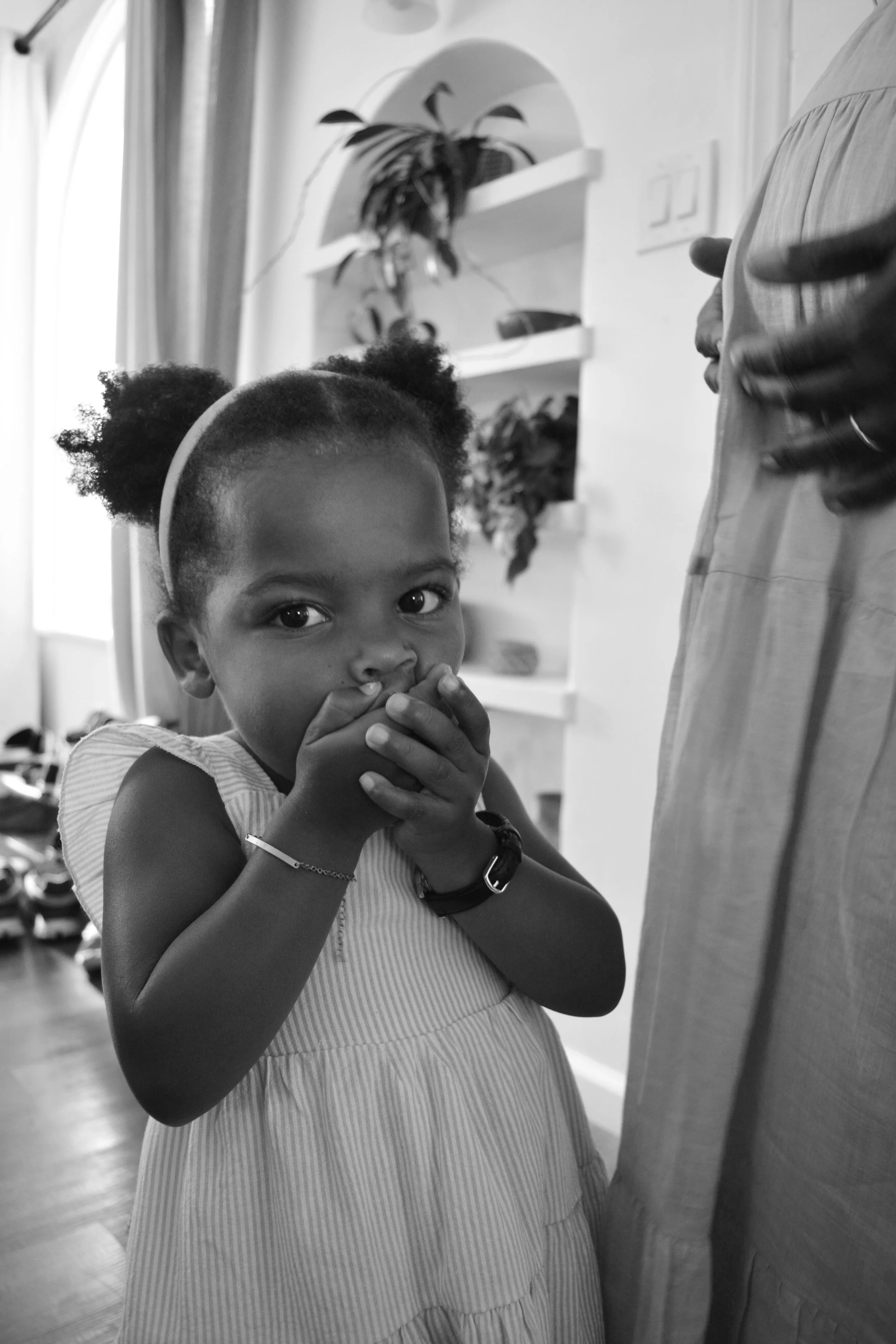 A young girl with curly hair in puffs, wearing a striped dress, holding her hands together near her face, standing indoors with a reflection and a plant in the background.