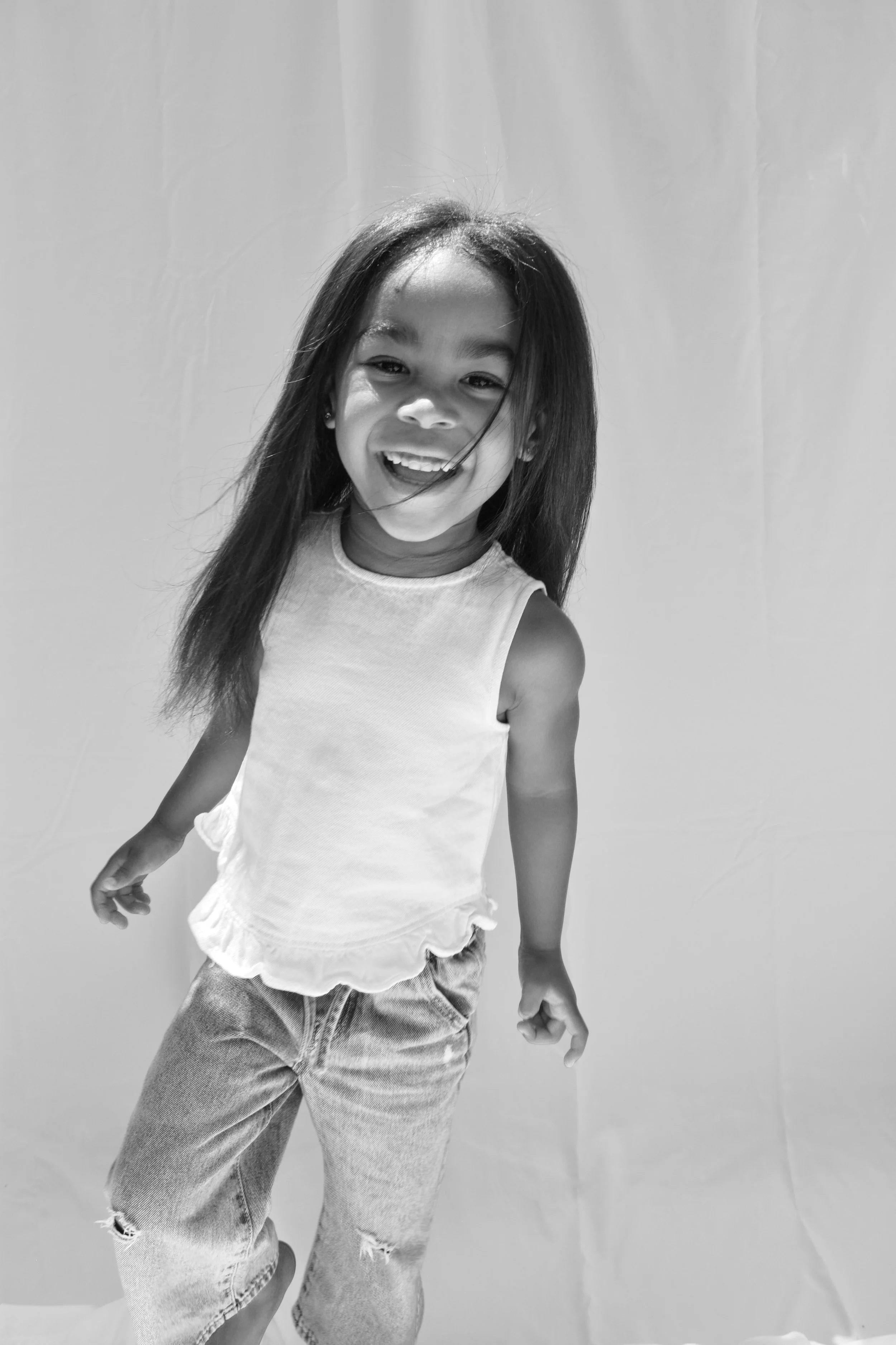 A young girl smiling and looking at the camera, wearing a sleeveless top and jeans, standing against a plain background.