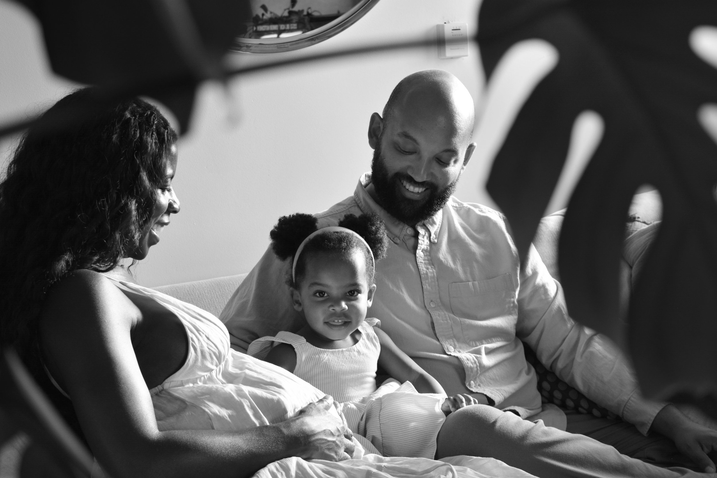 A family portrait in black and white showing a woman, a man, and a young girl sitting closely together on a couch, smiling and looking at each other.