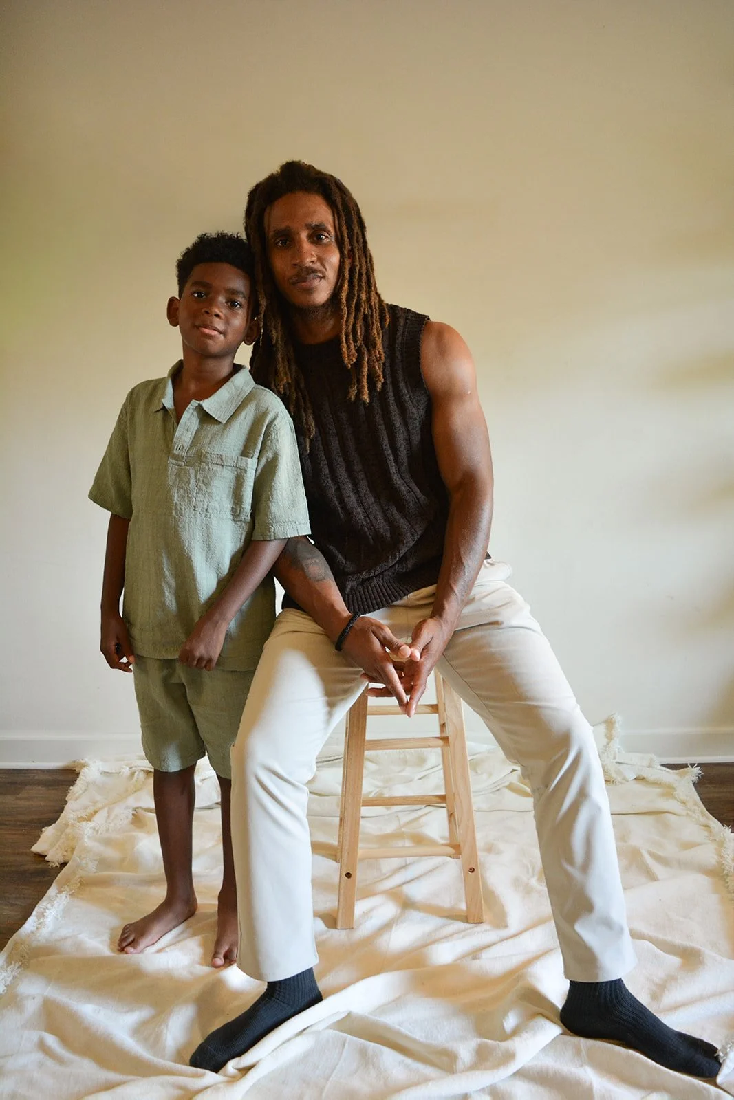 A man with dreadlocks and a young boy standing indoors against a plain wall, with a white cloth covering the floor.
