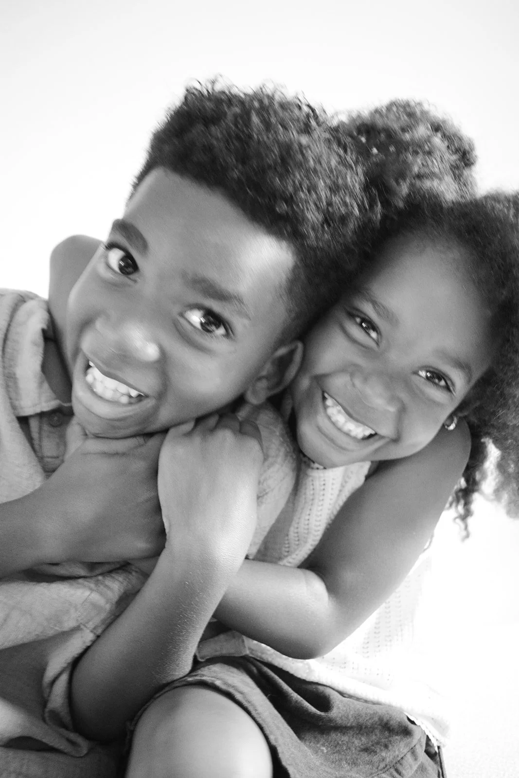 A black-and-white photo of two children, a boy and a girl, smiling and hugging. Both have natural hair and are dressed casually.