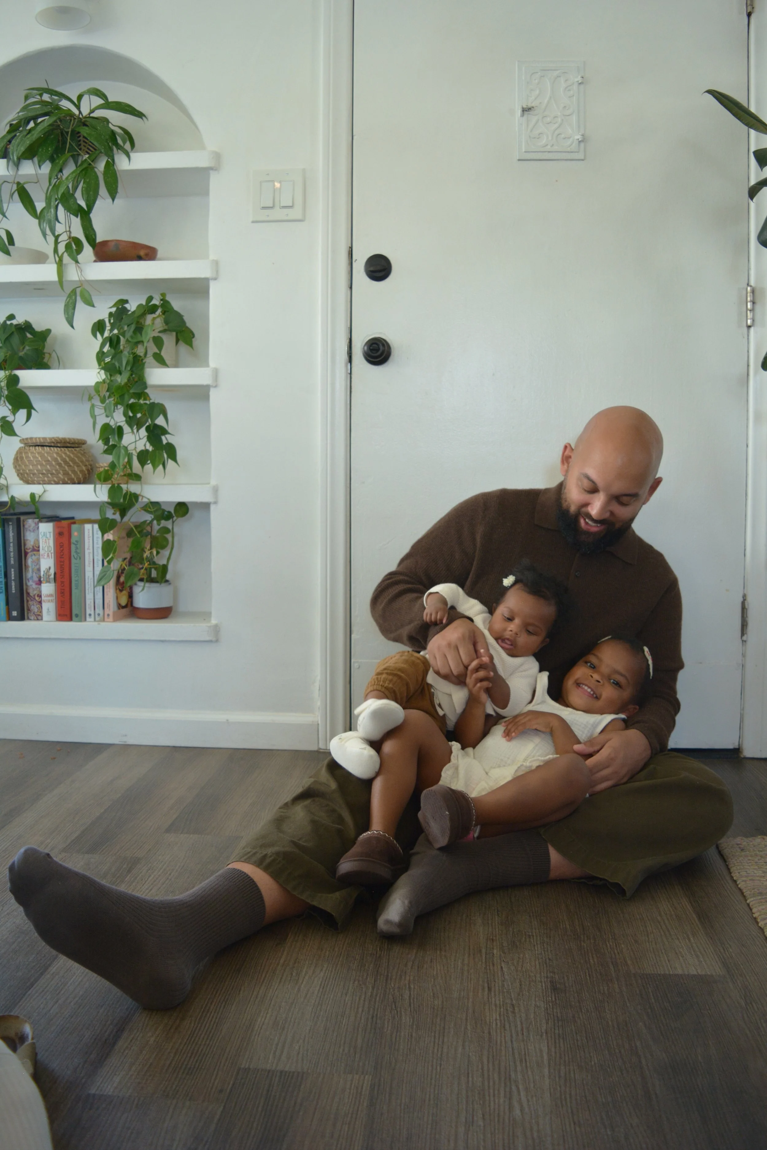 A man with a beard sitting on the floor in front of a white door, smiling and holding two young children, both wearing light-colored dresses, in a room with a bookshelf and green plants.